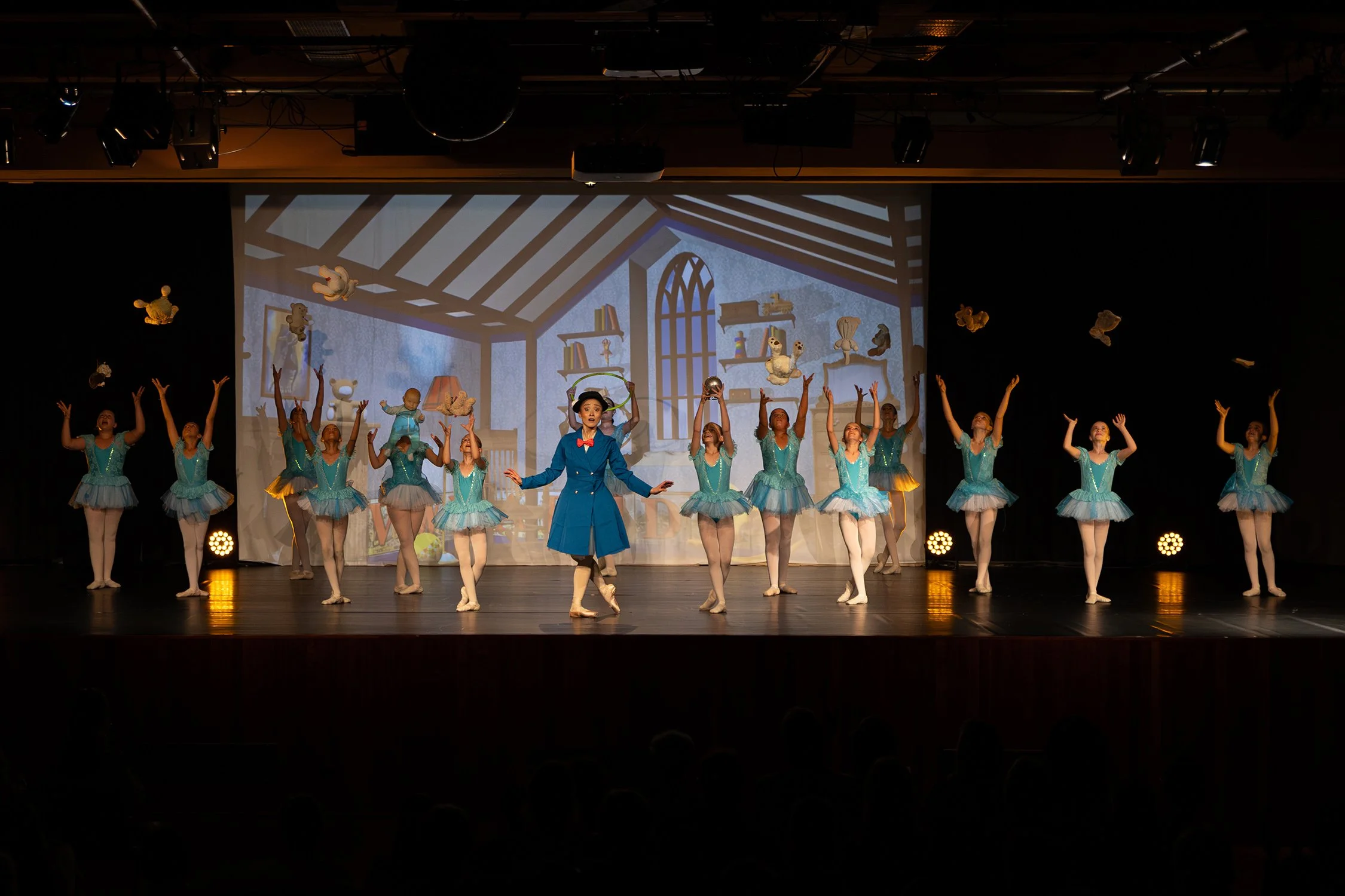 Des danseurs en tutu bleu exécutent une chorégraphie sur scène, avec un décor de dessin animé en arrière-plan représentant une pièce avec des livres et des jouets.