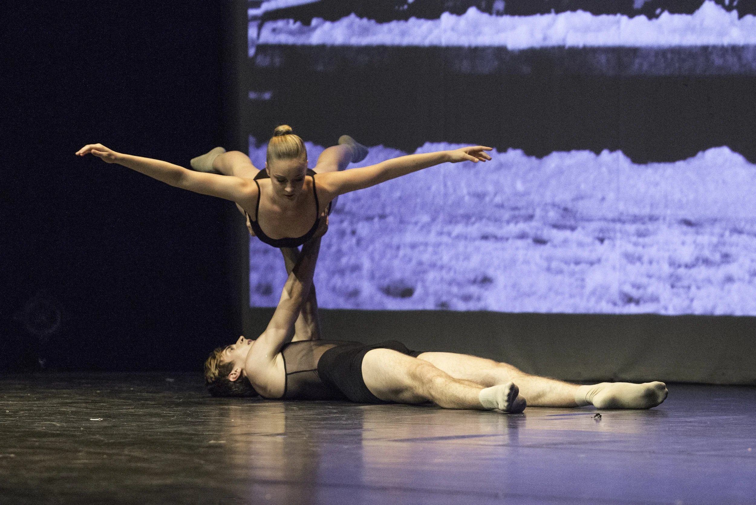 Un danseur homme allongé sur le sol, soutenant une danseuse femme qui est en équilibre au-dessus de lui, dans une scène de danse théâtrale ou ballet, avec un fond de projection d'images en noir et blanc.