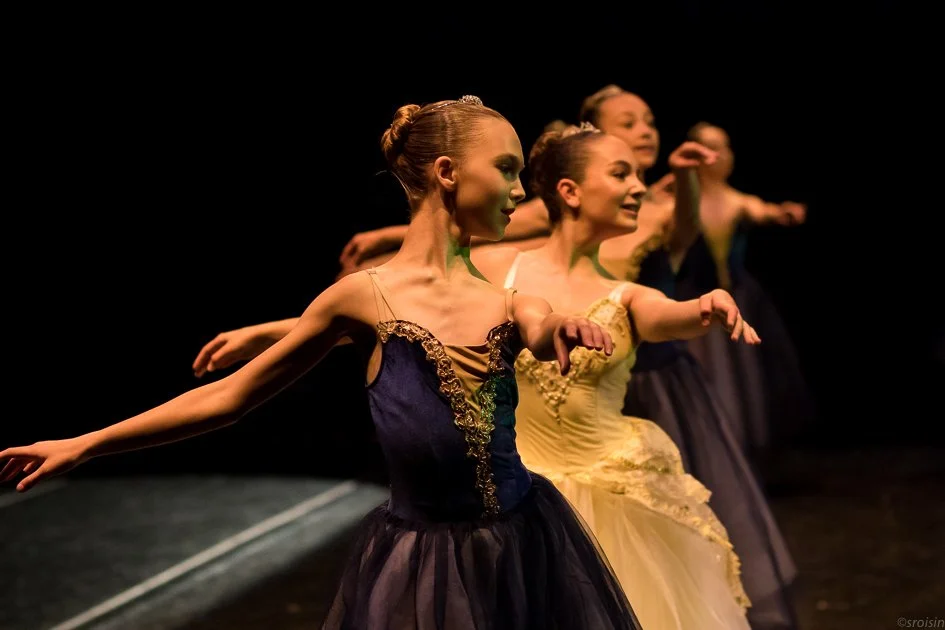Quatre ballerines en costume de scène exécutant une pirouette sur scène dans un éclairage sombre.