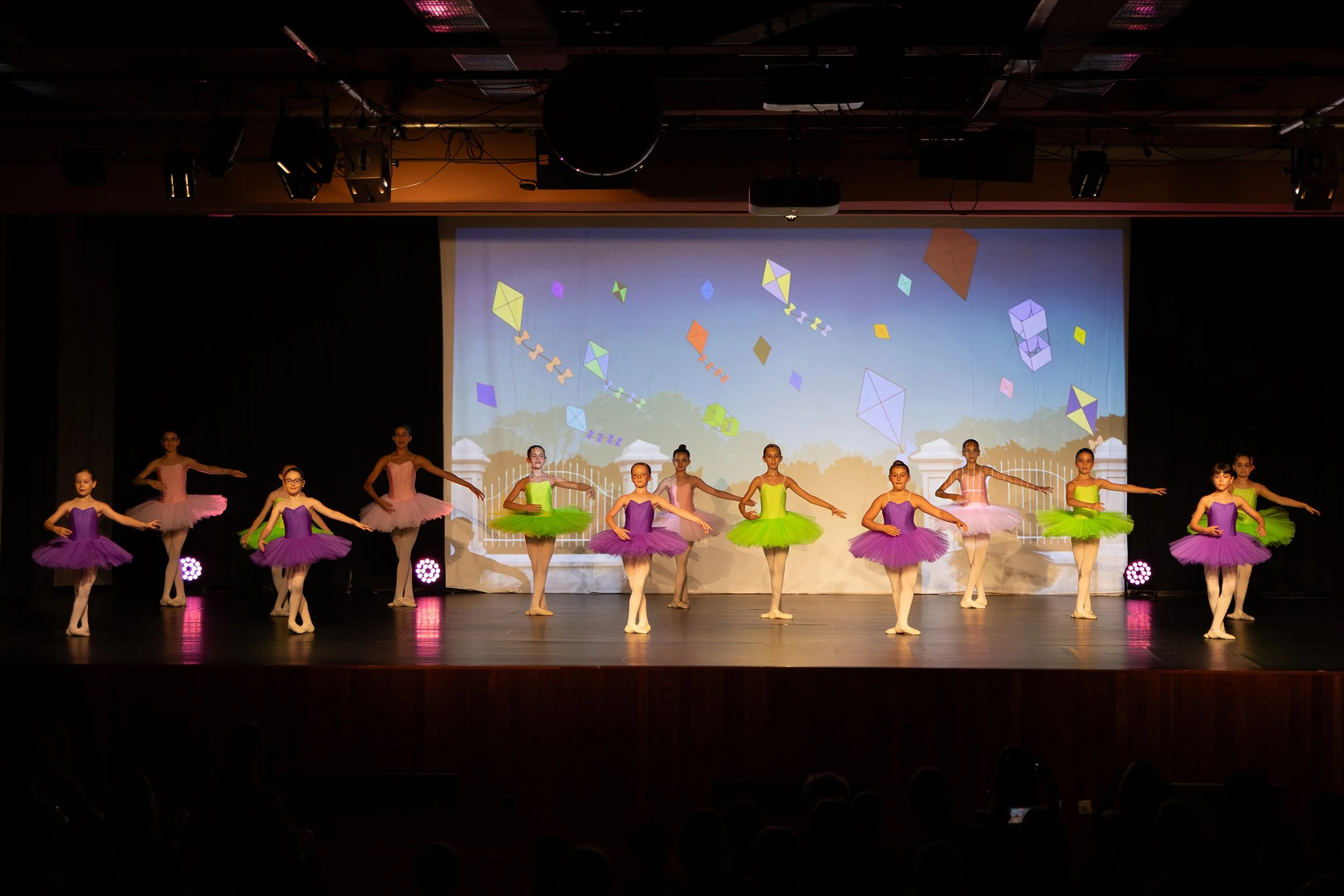 Jeunes ballerines en costume coloré, dansant sur scène lors d'un spectacle de ballet, avec un fond illustré de cerfs-volants.