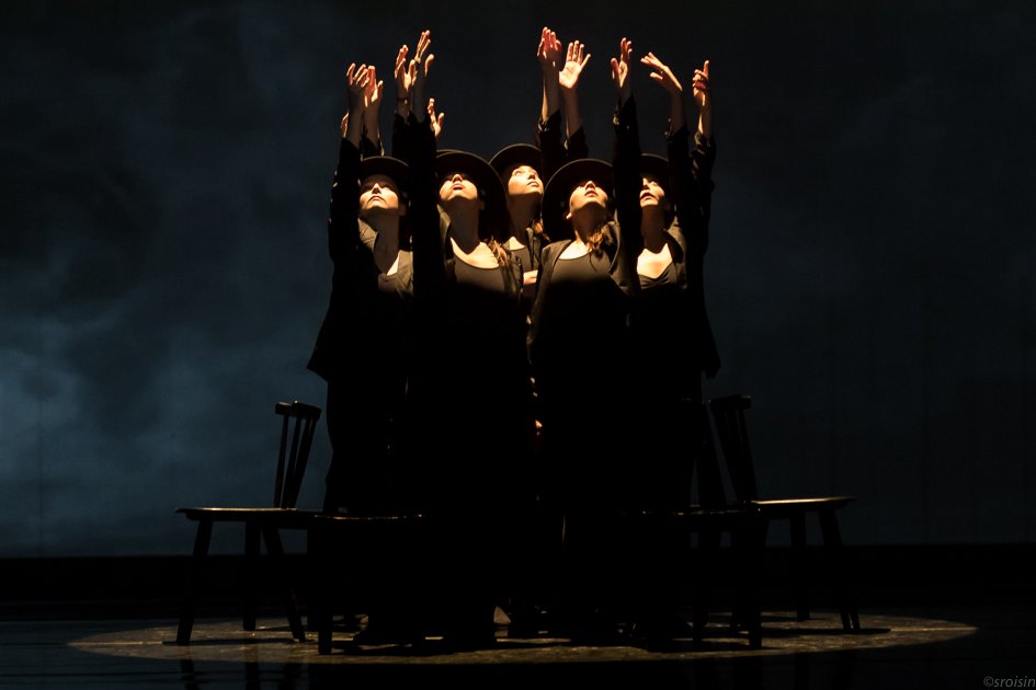 Cinq femmes habillées en noir, portant des chapeaux, levant leurs bras vers le haut sur scène dans une mise en scène dramatique.