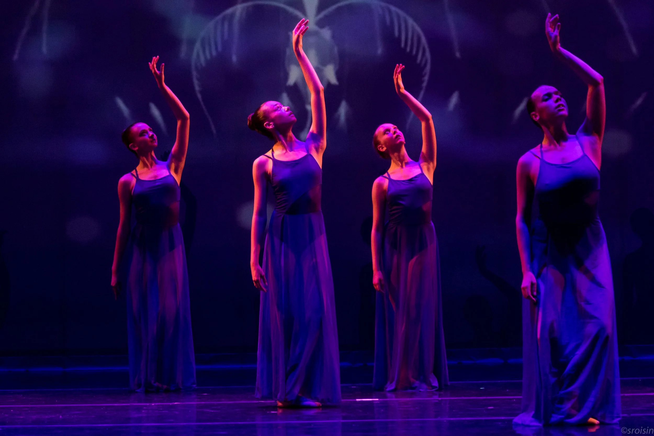 Quatre femmes ballerines en costume de scène violet exécutant une pose de ballet sur scène sombre