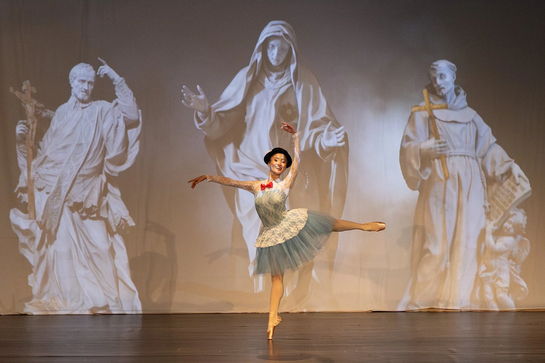 Une ballerine en tutu joue sur scène devant un fond projecté représentant des figures religieuses en blanc et gris.