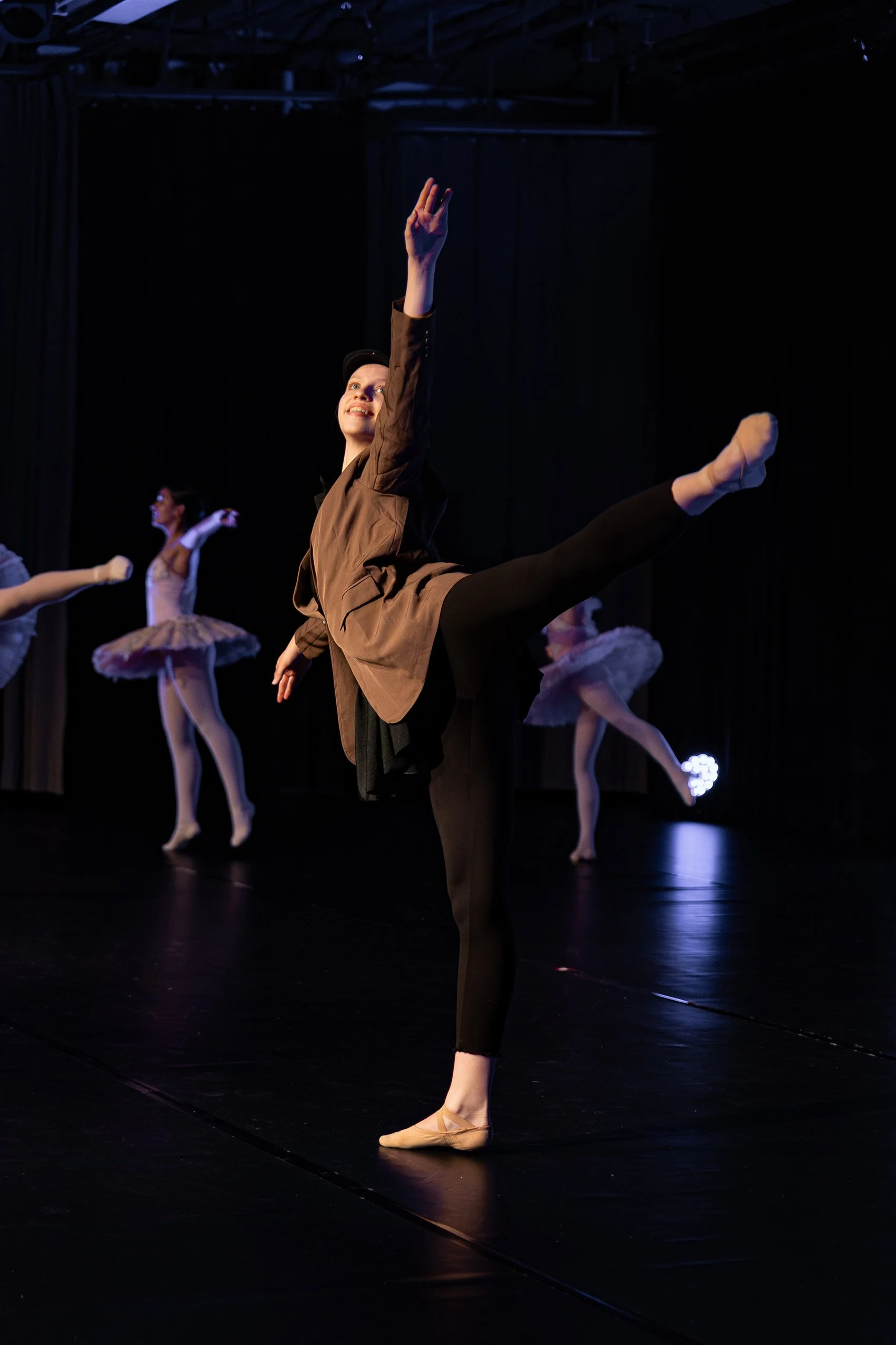 Des danseurs ballerines en costume de spectacle dans un studio de danse. La danseuse au premier plan exécute une pose en équilibre sur une jambe, avec l'autre jambe levée et étendue, dans une performance de ballet classique.
