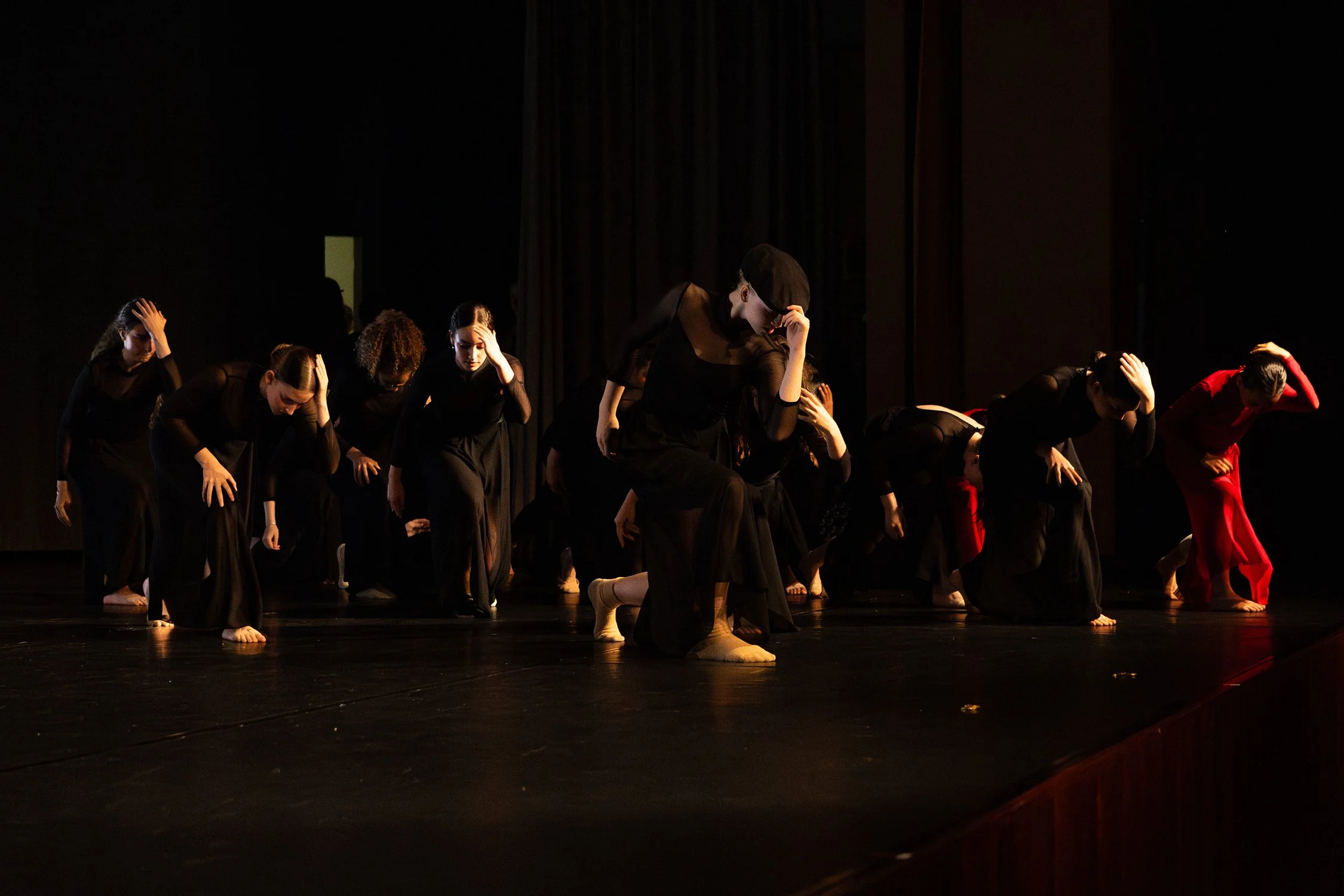 Groupe de danseurs sur scène, habillés en noir, effectuant une performance avec une position penchée, certains avec une main sur la tête, à l'avant-plan un danseur en bonnet noir avec une touche de rouge à l'arrière.