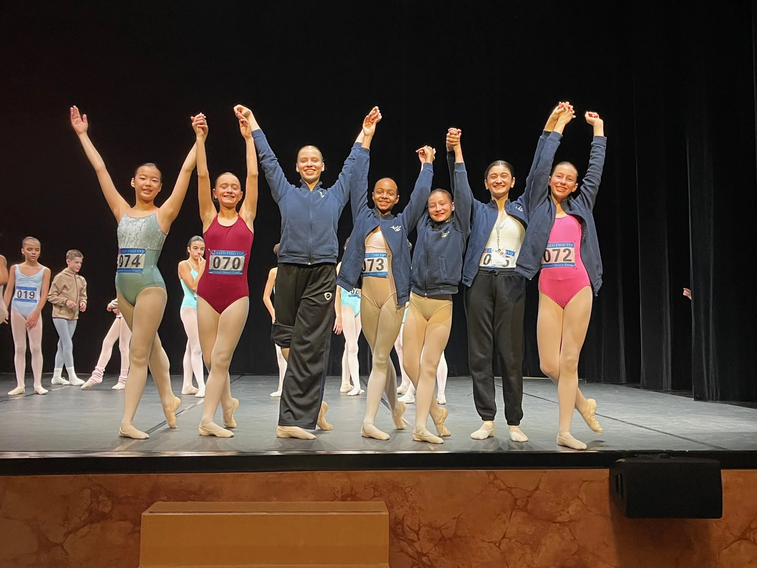 Groupe de jeunes danseurs en costume de ballet sur scène, certains portant des vestes, célébrant la réussite, avec d'autres danseurs en arrière-plan.