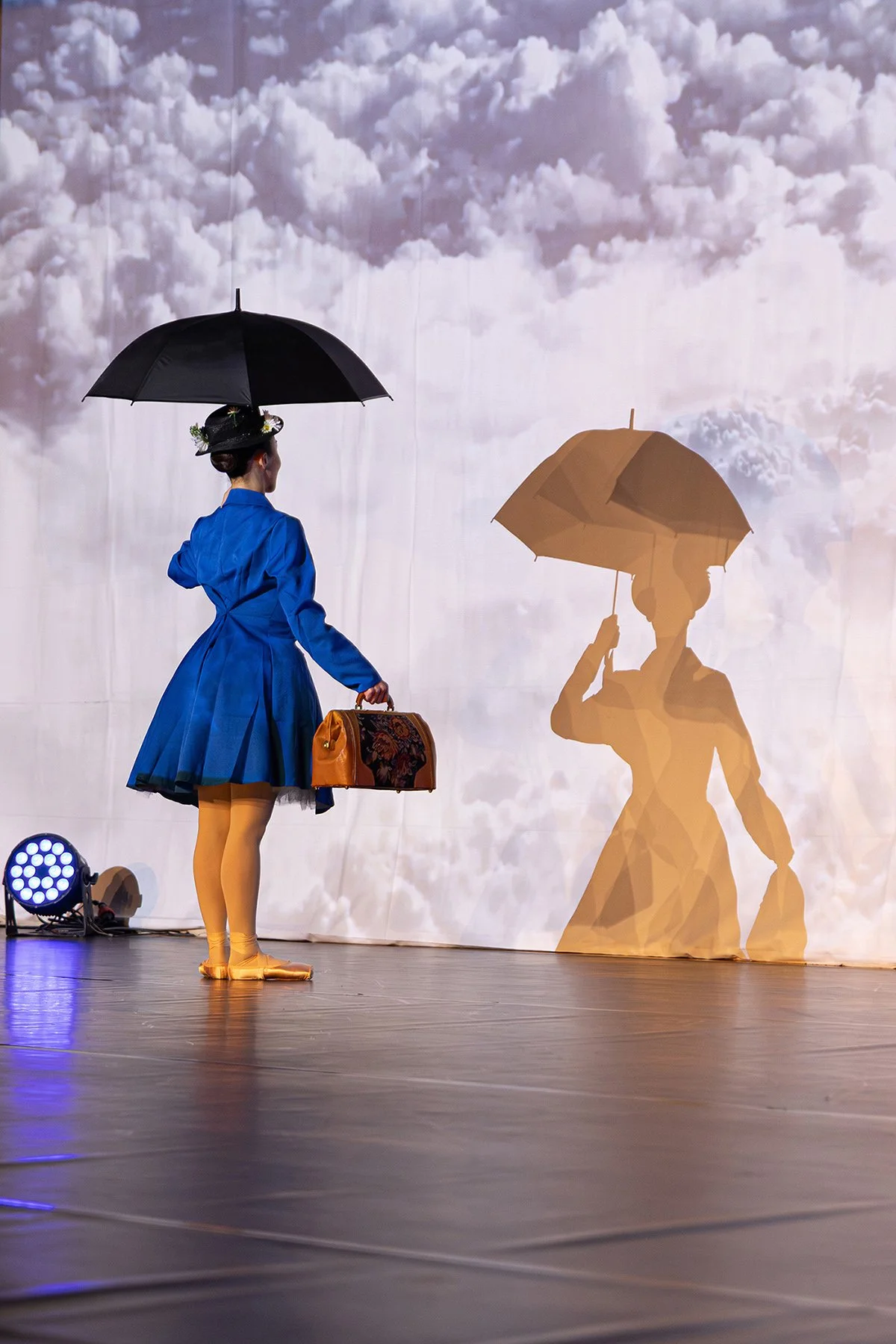 Une femme en costume bleu, portant un chapeau orné de fleurs, tient un parapluie noir et un sac orange, se tenant devant un fond de nuages projetant son ombre sur un rideau.