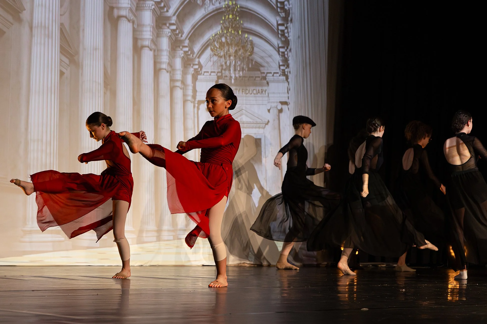 Jeunes danseurs en costumes noirs et rouges exécutant une performance de danse classique sur scène, avec un décor d'architecture classique en arrière-plan.