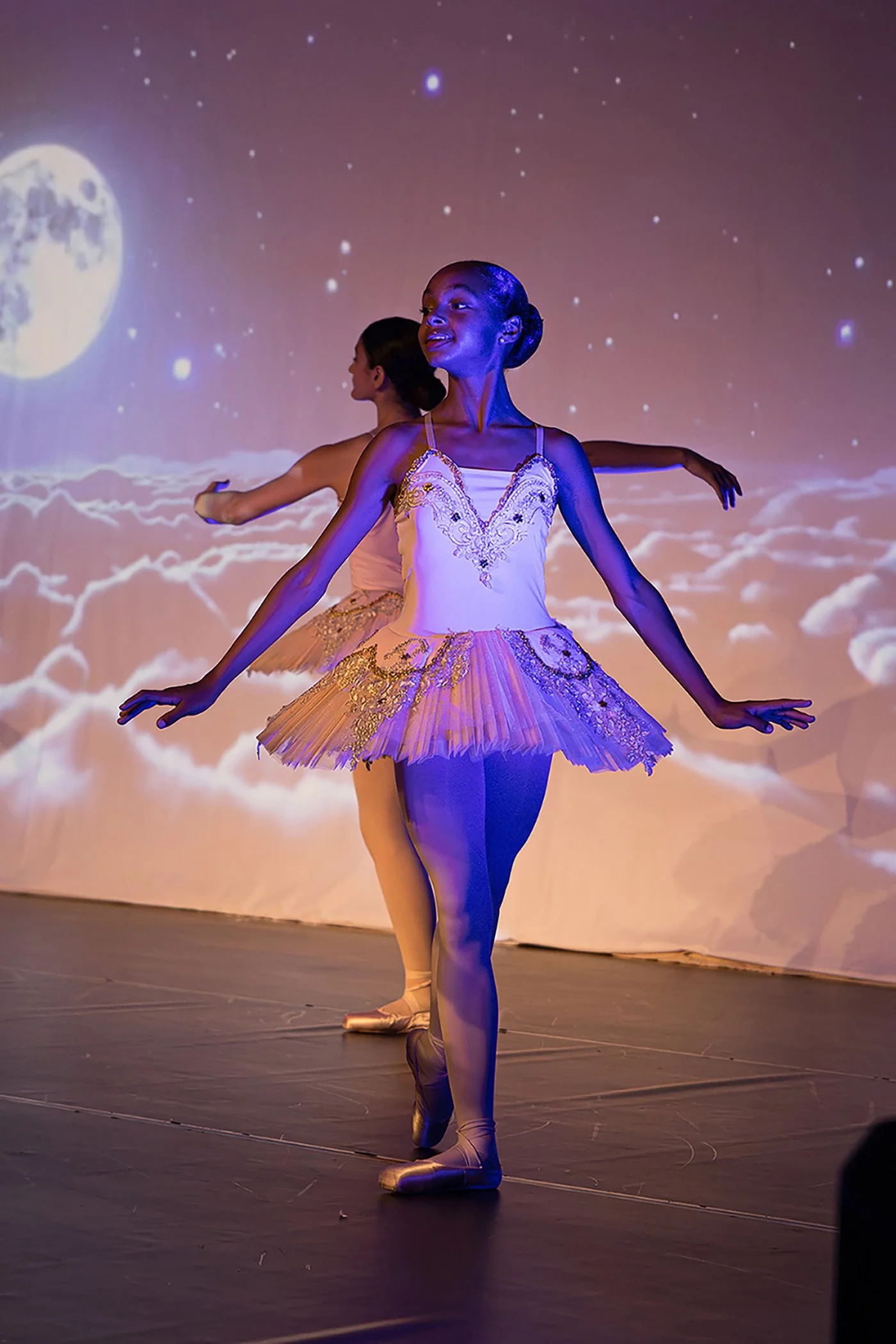 Deux danseuses classiques en tutu, dansant sur scène avec un fond d'écran d'une lune et des étoiles, éclairée par des lumières bleues et roses.