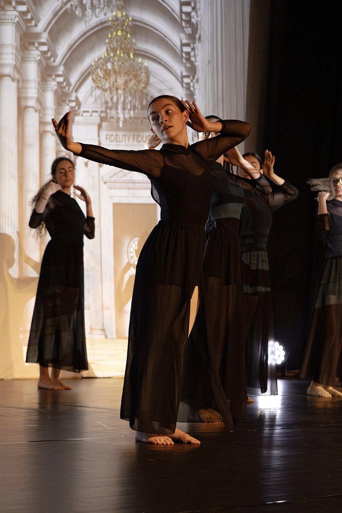 Groupe de danseuses en costume noir, en entraînement ou en performance sur scène, avec décor intérieur classique et chandelier en arrière-plan.