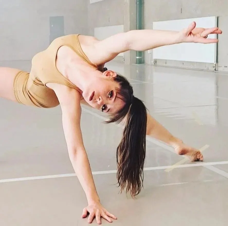 Une femme réalise une pose de danse contemporaine dans une salle très lumineuse, elle est en position de planche avec une main au sol et une jambe levée.