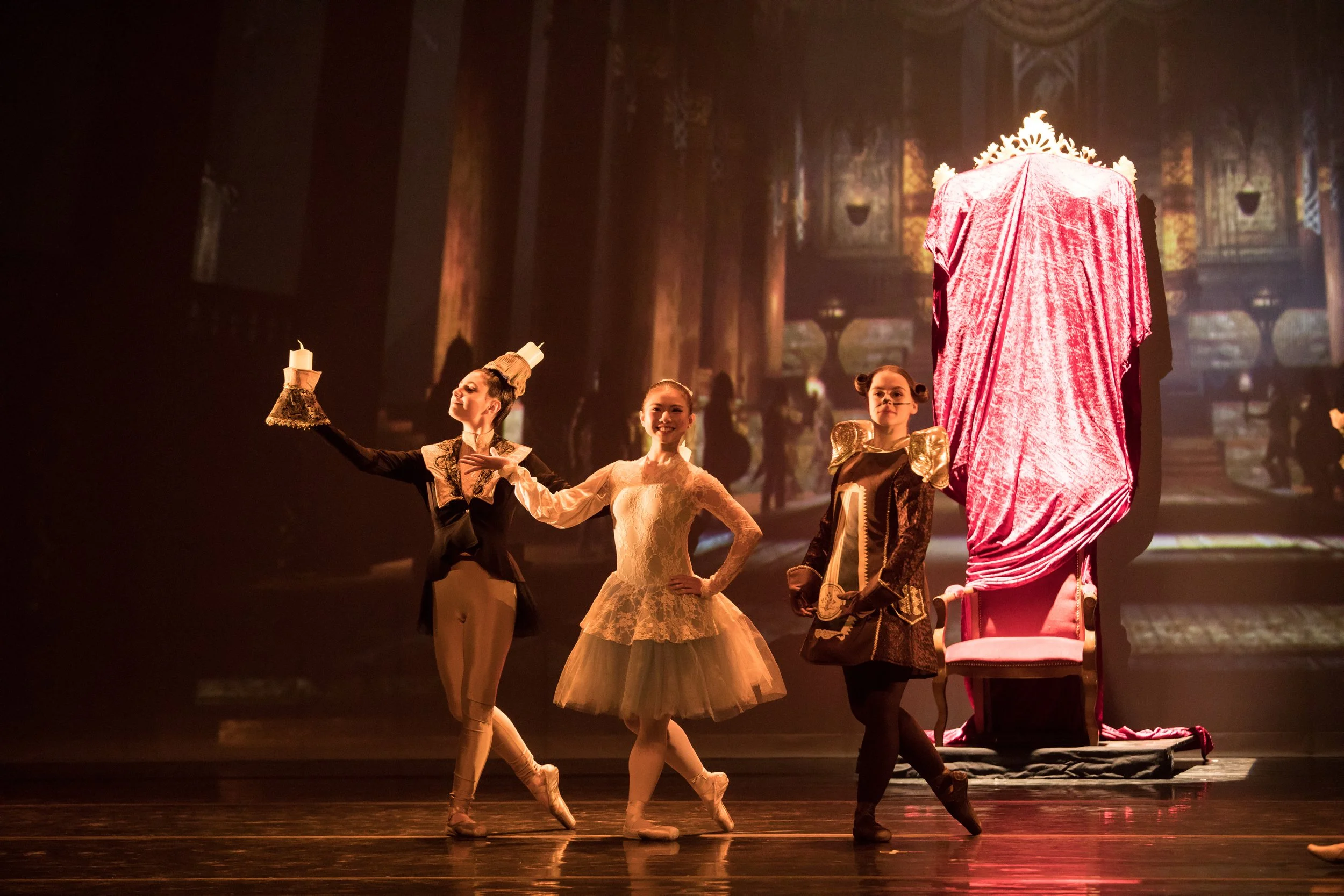 Trois ballerines en costumes de ballet posent devant un décor de théâtre avec un trône drapé de velours rouge.