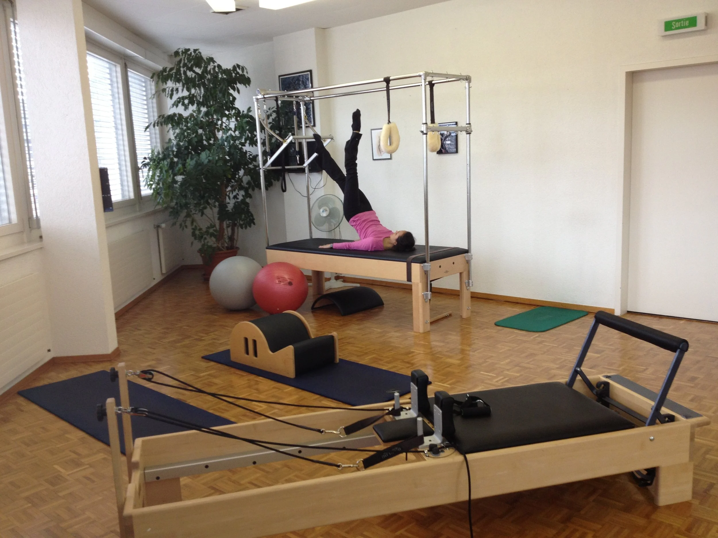 Une femme en séance de rééducation ou de fitness dans une salle avec divers équipements, notamment un appareil à cadre pour exercices, ballons de gymnastique, tapis, et autres accessoires. Pilates à Chêne-Bourg.