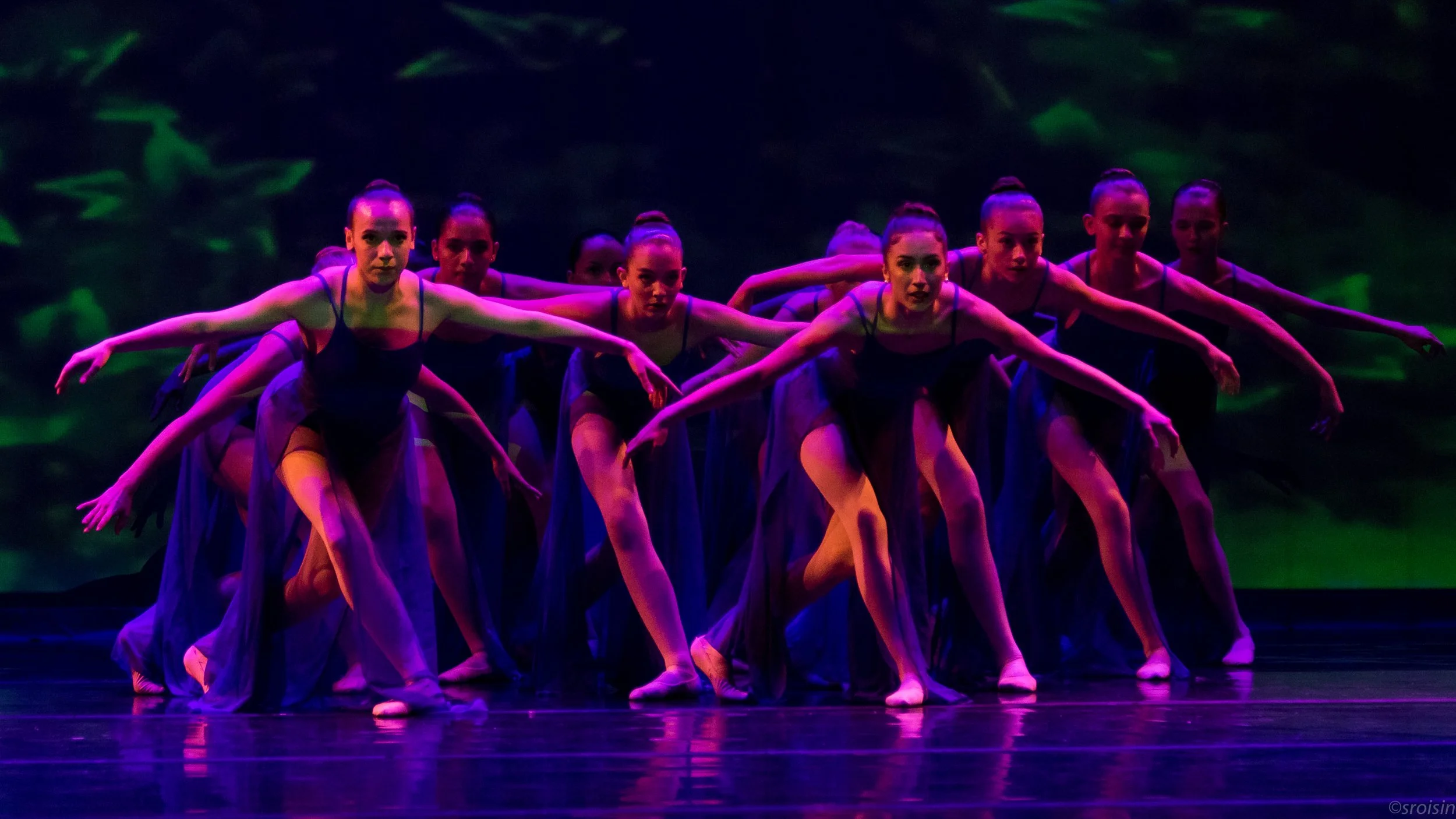 Un groupe de danseuses classiques en costume noir effectuant une performance sur scène, éclairée de lumières violettes et vertes.