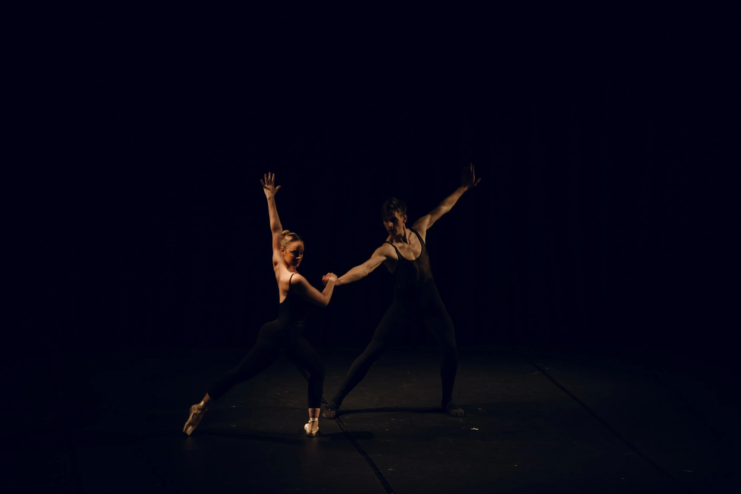 Deux danseurs en tenues noires dans une scène sombre, exécutant une pose de danse moderne, la femme sur pointe et l'homme tenant sa main.