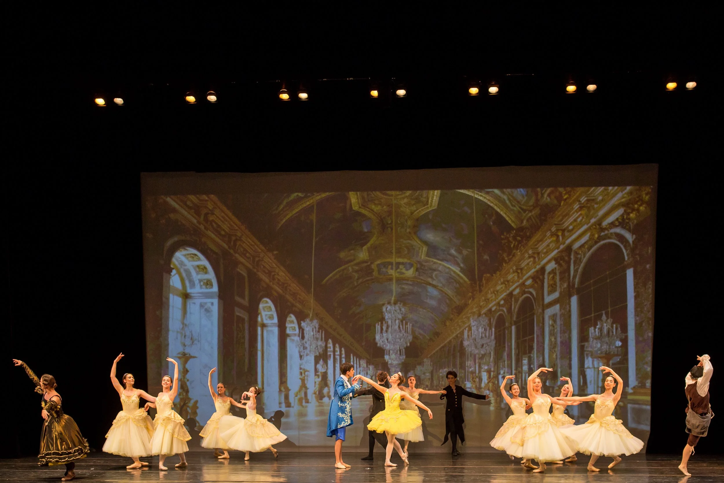 Une scène de ballet avec plusieurs danseurs en costumes élaborés, dont certains en tutu jaune, sur un fond d'écran représentant un intérieur somptueux avec chandeliers et plafonds ornés.