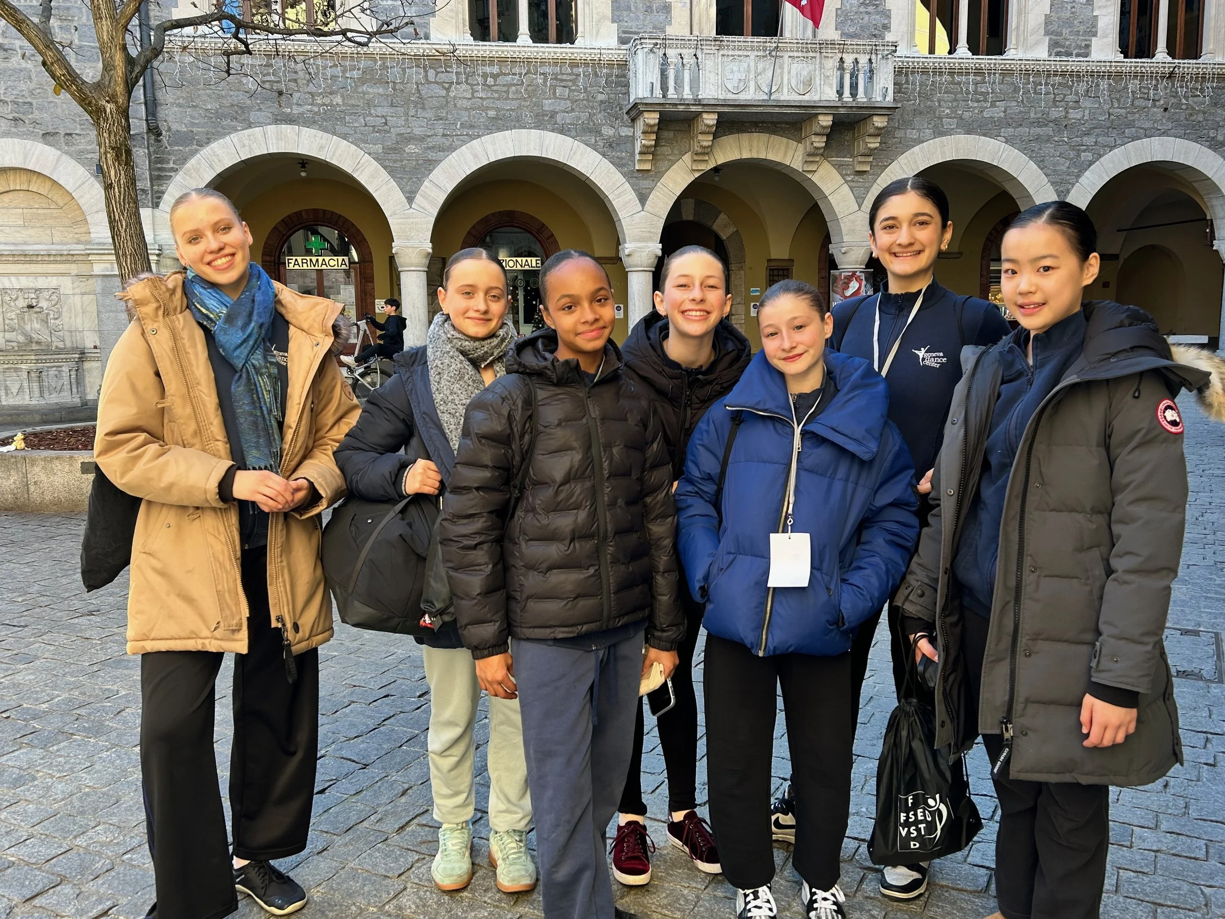 Groupe de sept jeunes filles posant pour une photo devant un bâtiment en pierre avec des arches, certains portant des sacs à dos et des vêtements chauds, dans un espace public en extérieur.