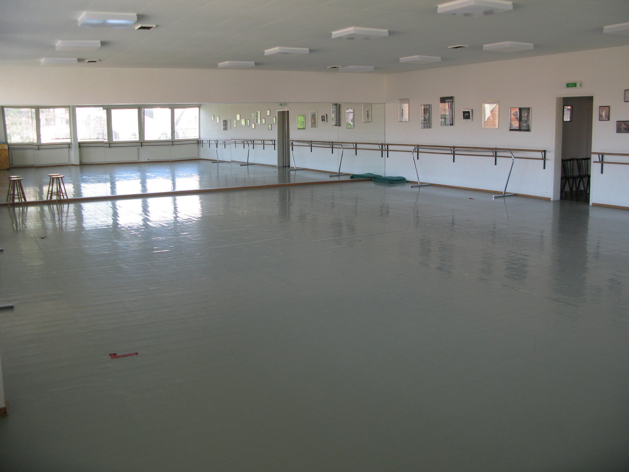 Une grande salle de danse vide, des miroirs sur le mur et des barres de ballet, éclairée par de grandes fenêtres et des plafonds blancs. Geneva dance center.
