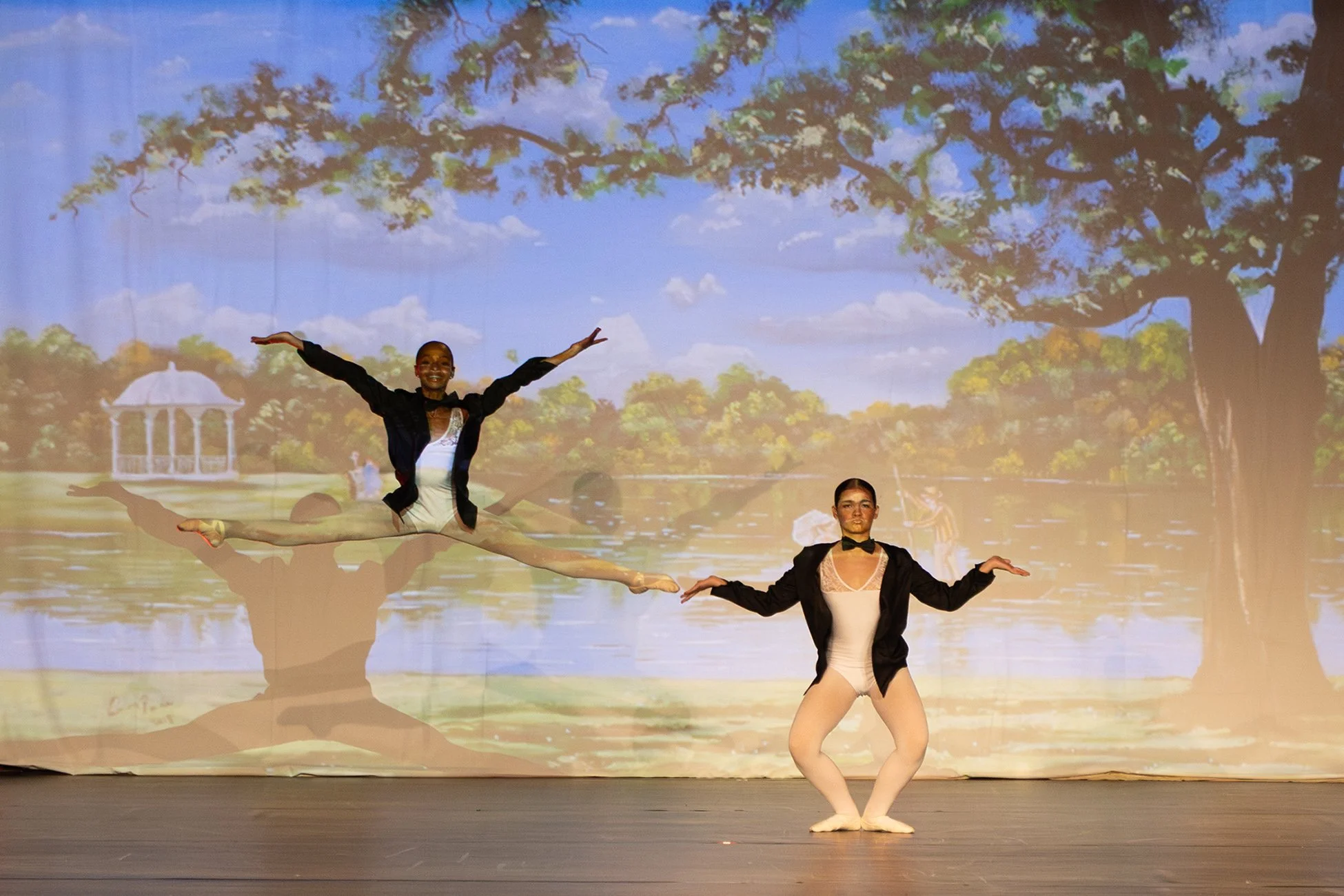 Deux danseurs sur scène, un levant une jambe en l'air et l'autre positionnée au sol, avec une toile de fond représentant un parc avec un arbre et un pavillon, lors d'une performance de danse.
