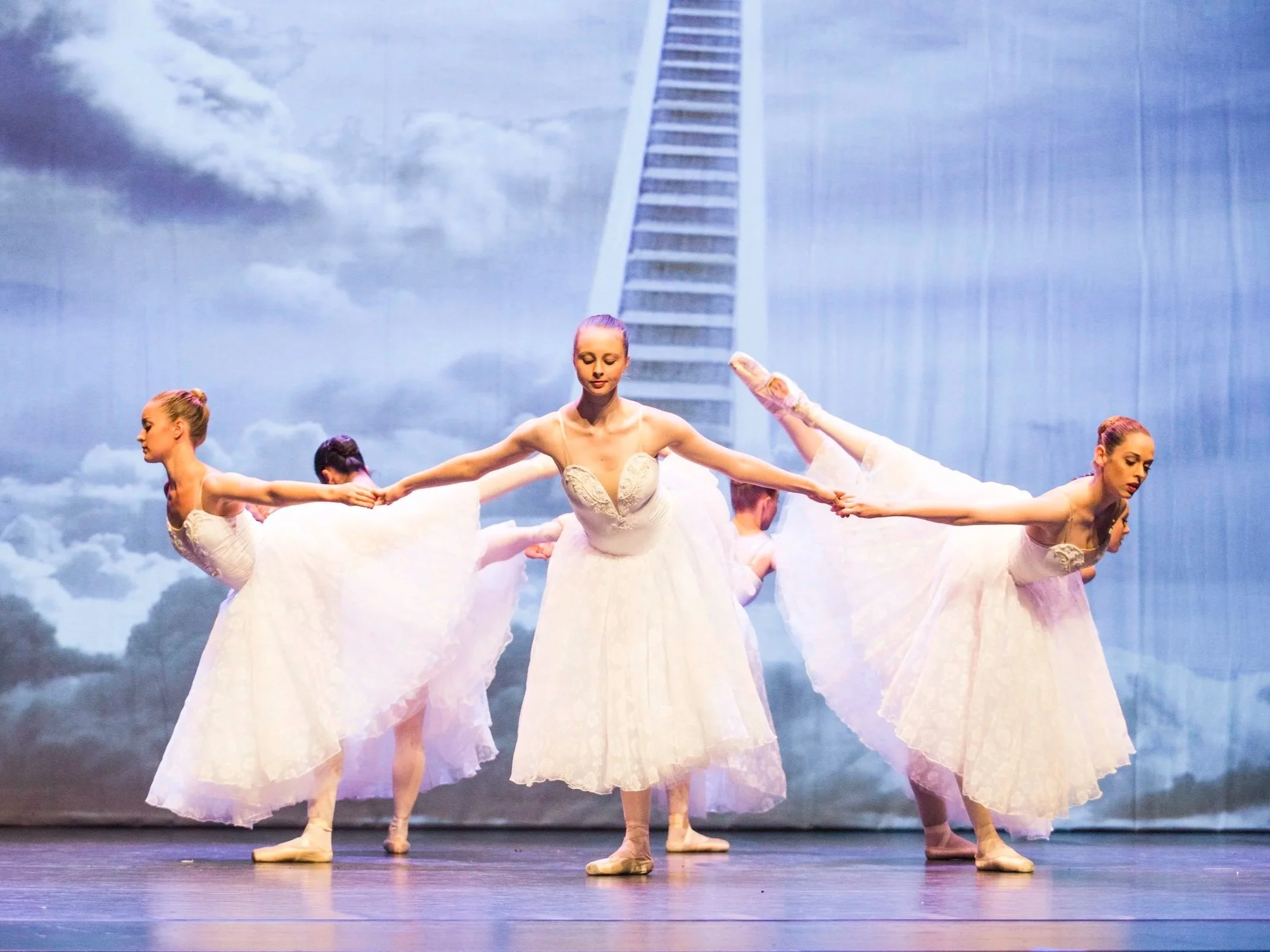 Groupe de ballerines en costume blanc sur scène avec un arrière-plan représentant un ciel nuageux et une échelle géante.