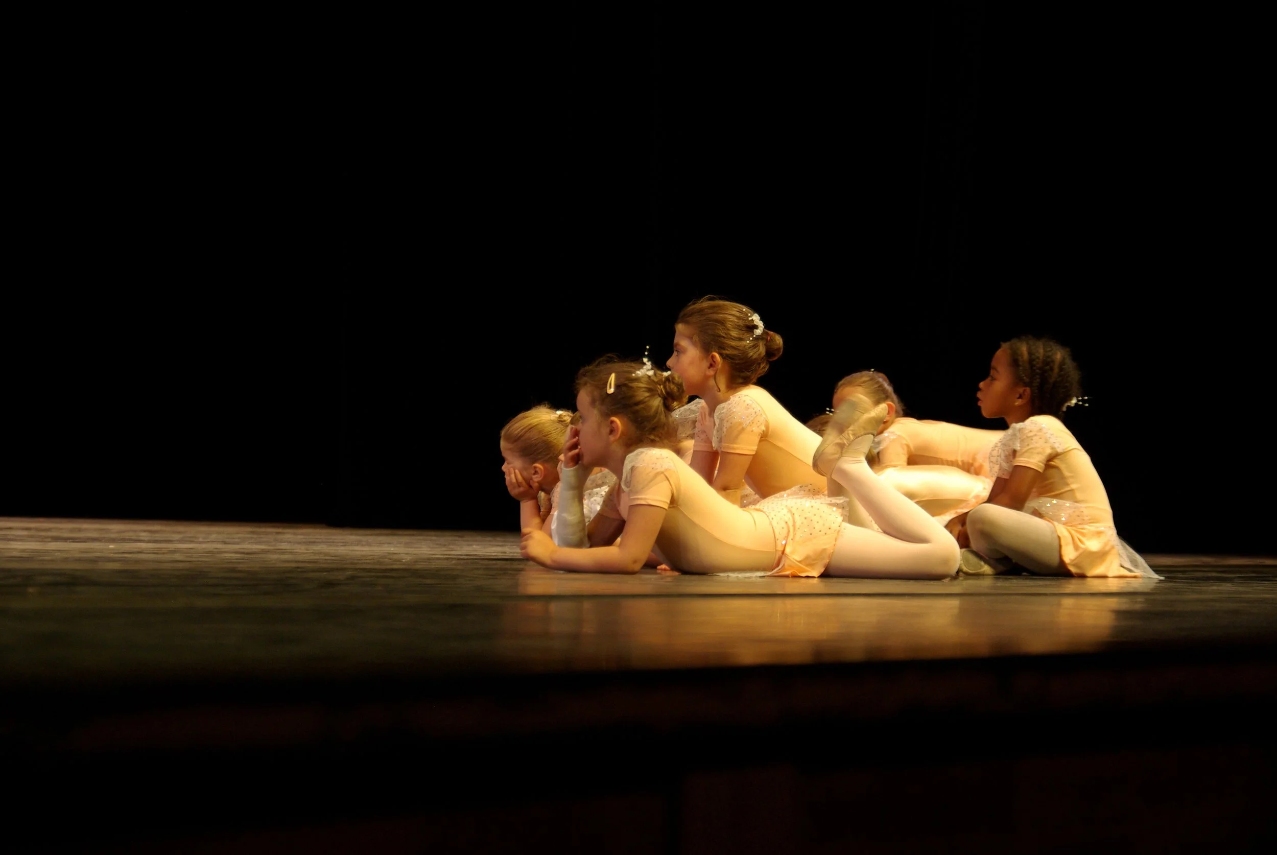 Cinq jeunes filles en costume de danse, allongées sur le sol d'une scène de théâtre, semblent attendre leur tour ou écouter une instruction, avec un fond noir.