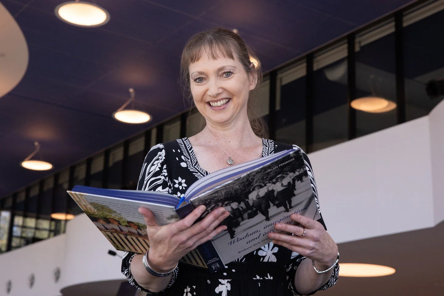 A woman with light skin and brown hair, smiling and reading a book in an indoor setting with round ceiling lights.