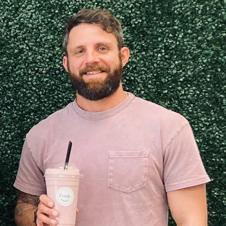 A man with a beard and curly hair smiling and holding a pink beverage in a clear cup with a black straw, standing in front of a green hedge background.