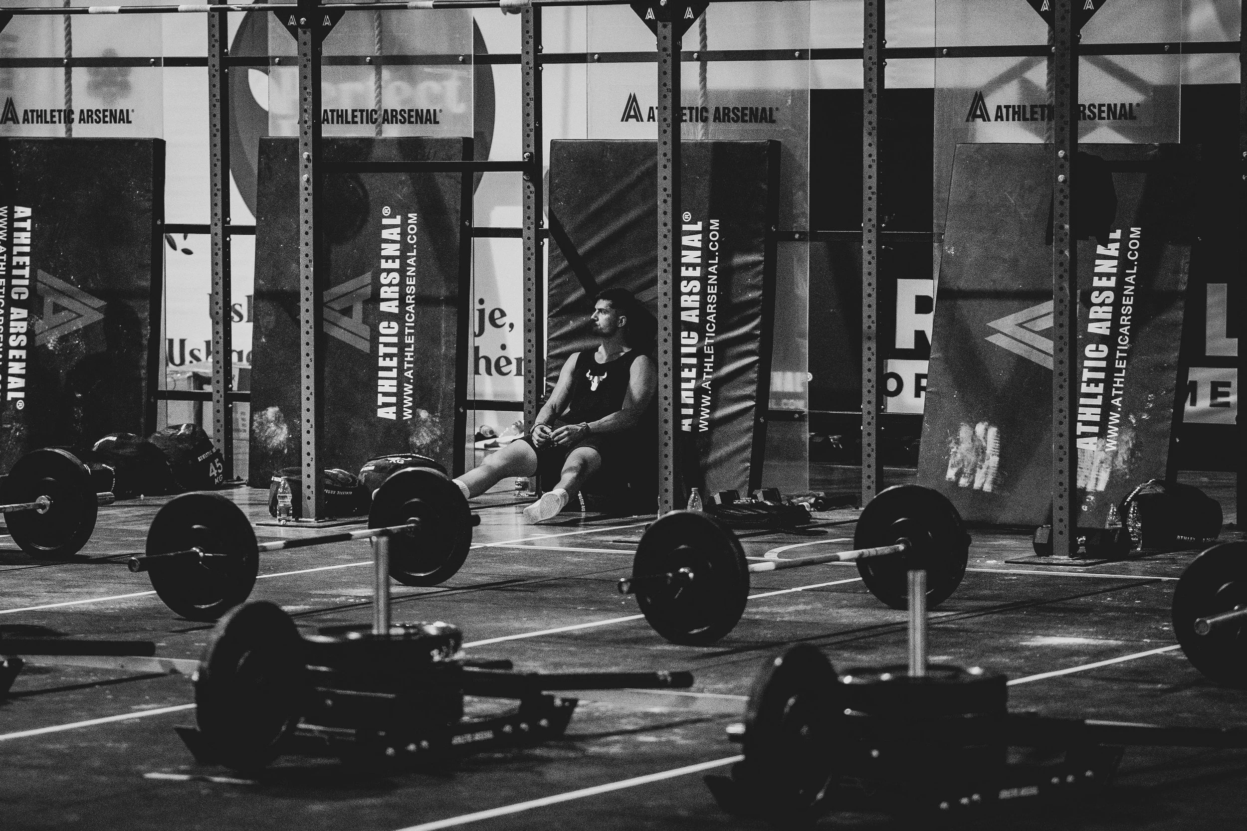A person sitting on the floor of a gym, surrounded by weightlifting equipment and racks, with banners displaying 'Athletic Arsenal' in the background.