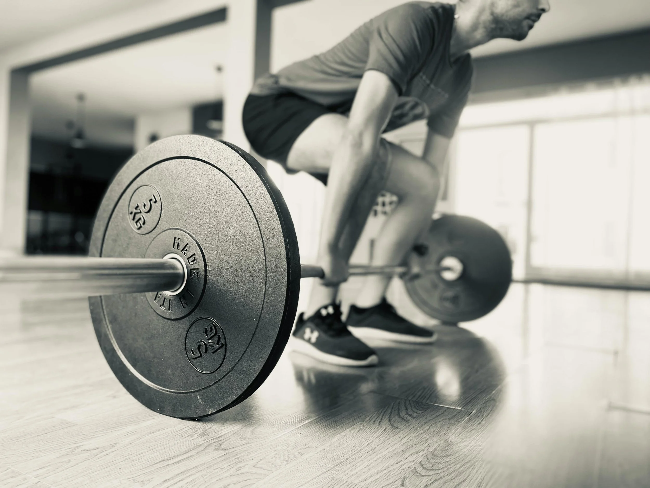 A man in athletic clothing is in the middle of performing a deadlift exercise at a gym, with a barbell loaded with 35 kg weights on the floor in front of him.