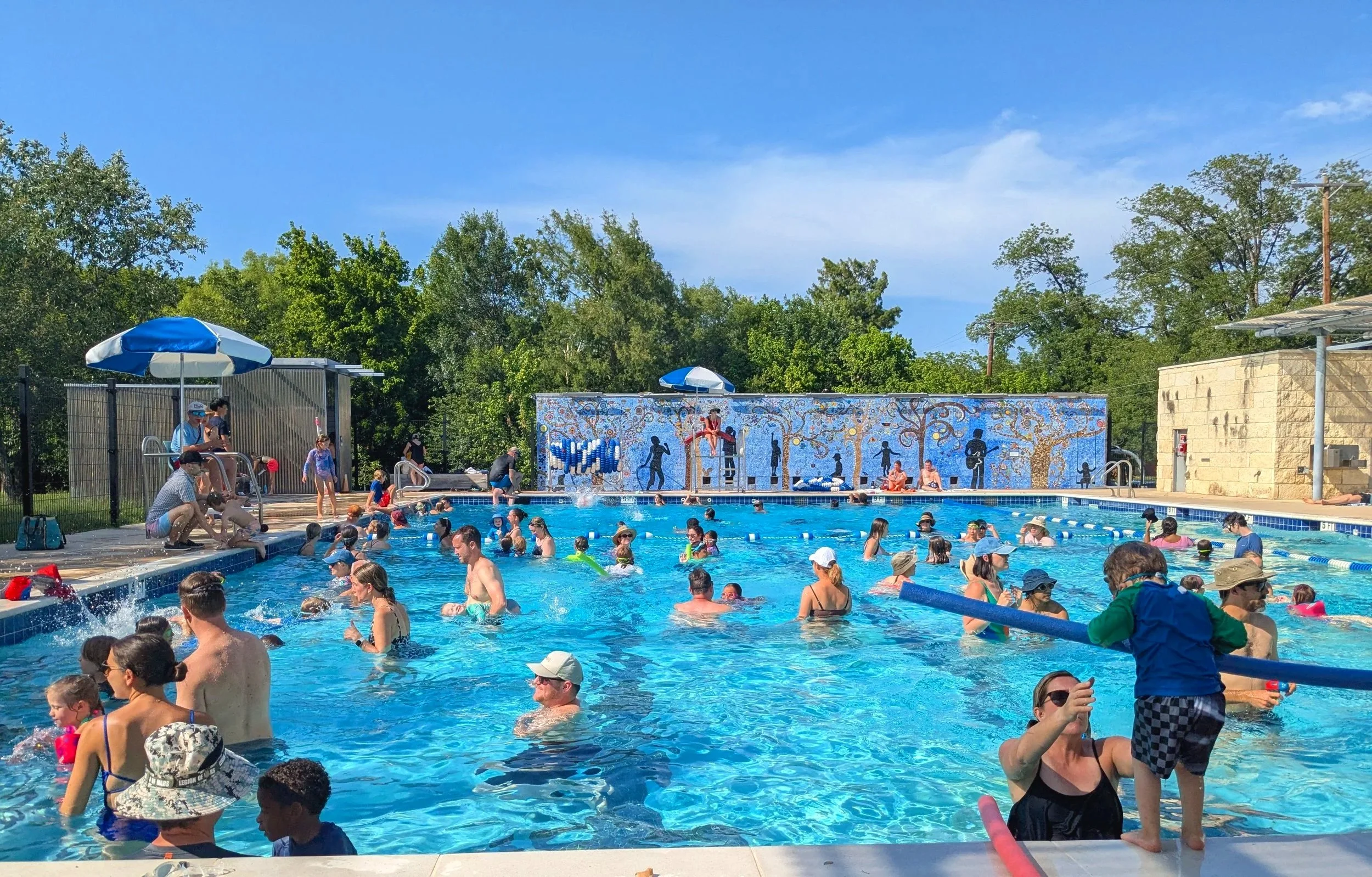 People swimming and relaxing at a busy outdoor public pool on a sunny day with blue sky, trees, and umbrellas.