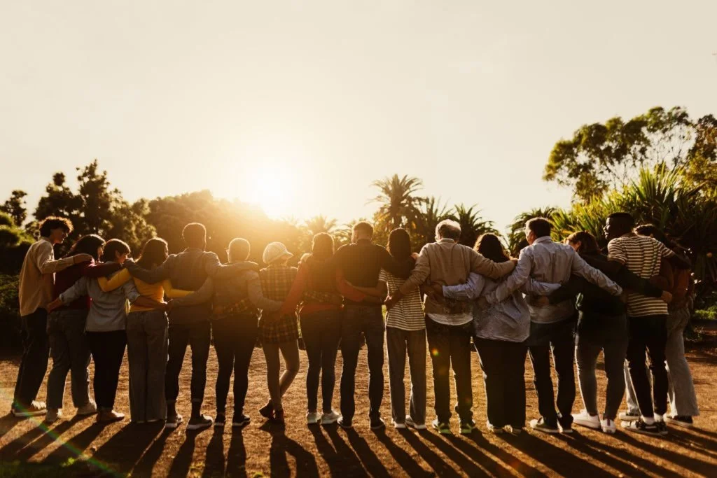 Group of diverse people standing outdoors at sunset, holding each other and forming a line, with shadows cast on the ground.