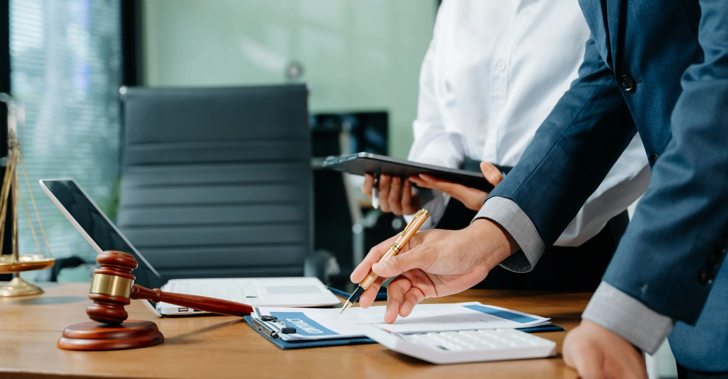 Business professionals discussing documents on a desk, with a gavel, laptop, and calculator in an office setting.