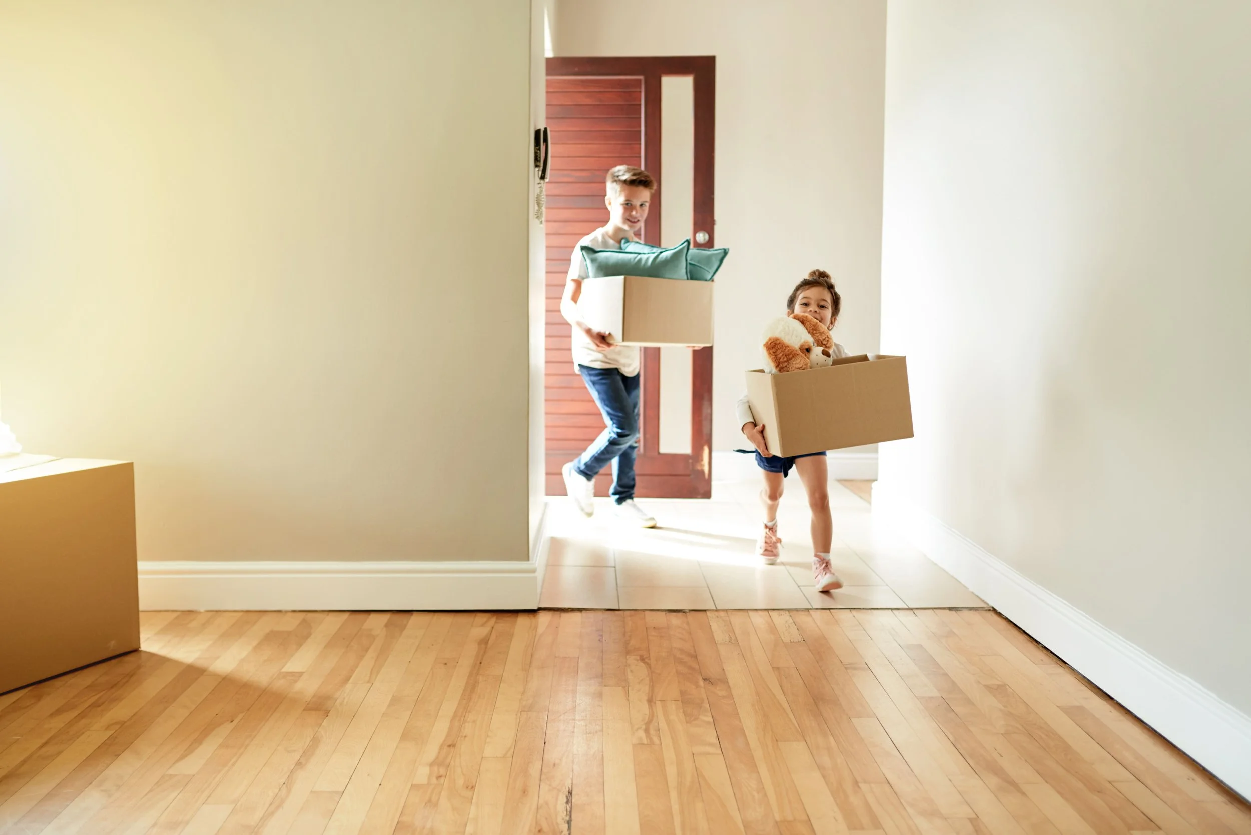 Two children, a boy and a girl, carrying boxes with stuffed animals inside, walking through a doorway into a room with wooden flooring and minimal decor, sunlight streaming in.
