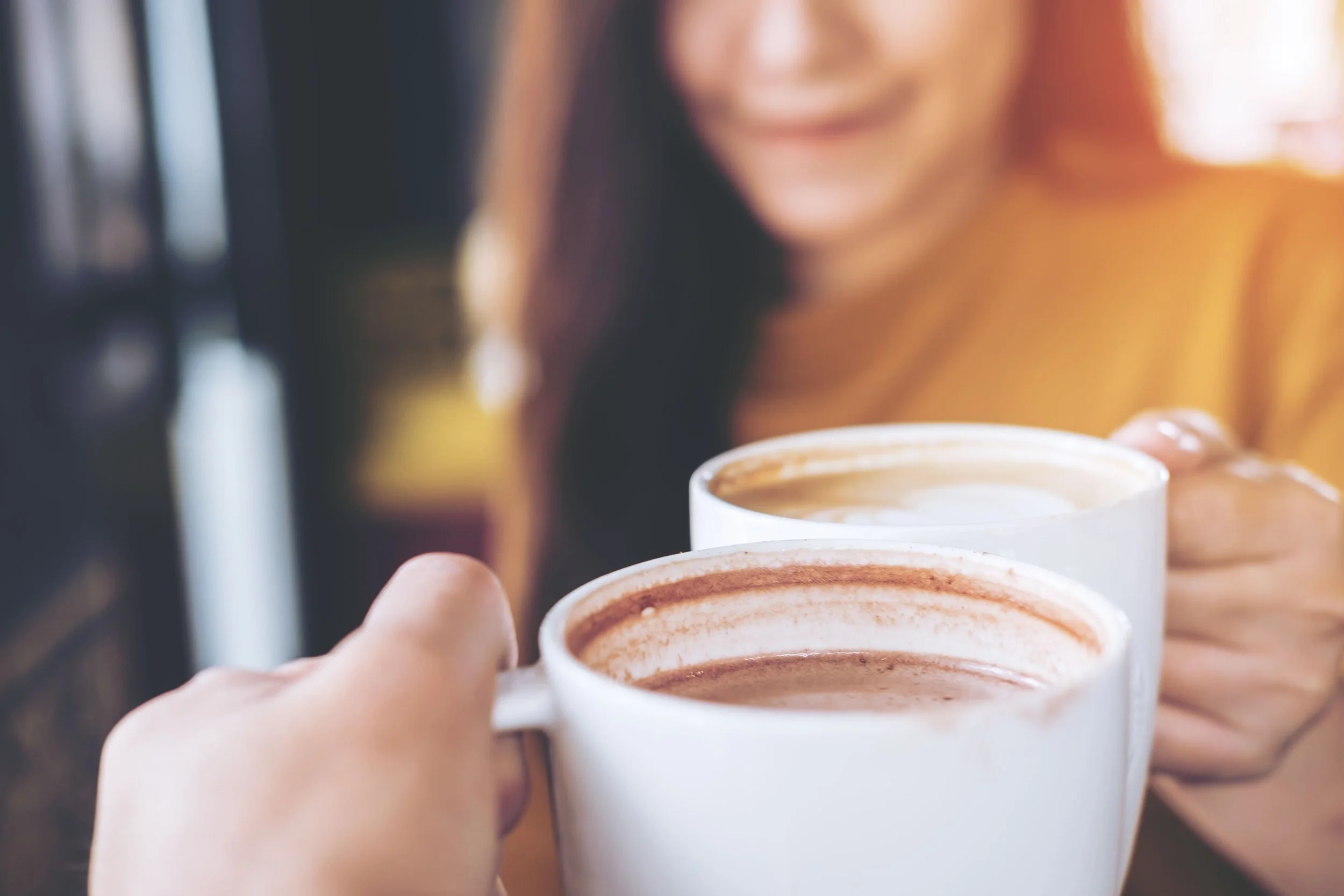 Two people clinking white coffee mugs filled with hot chocolate or coffee, with a smiling person in the background wearing a yellow top.