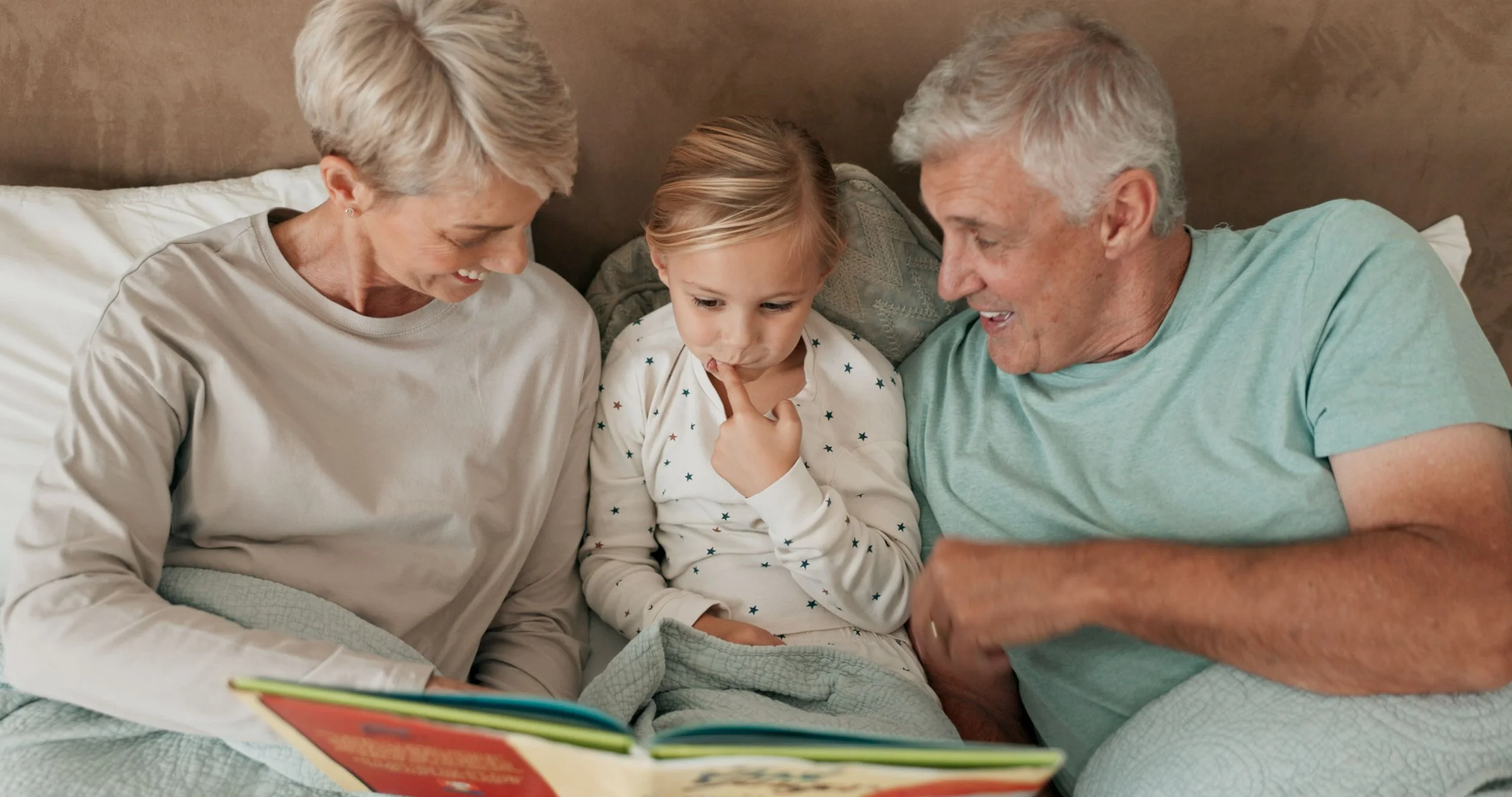 An elderly woman, a young girl, and an elderly man sitting together in bed, reading a book and smiling.