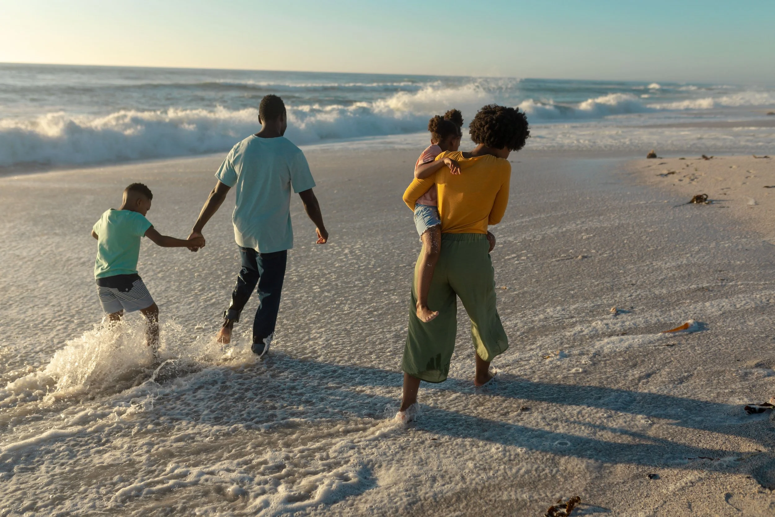 A family of five walking along the beach at sunset, with ocean waves in the background. The adults are carrying children, and the children are holding hands.
