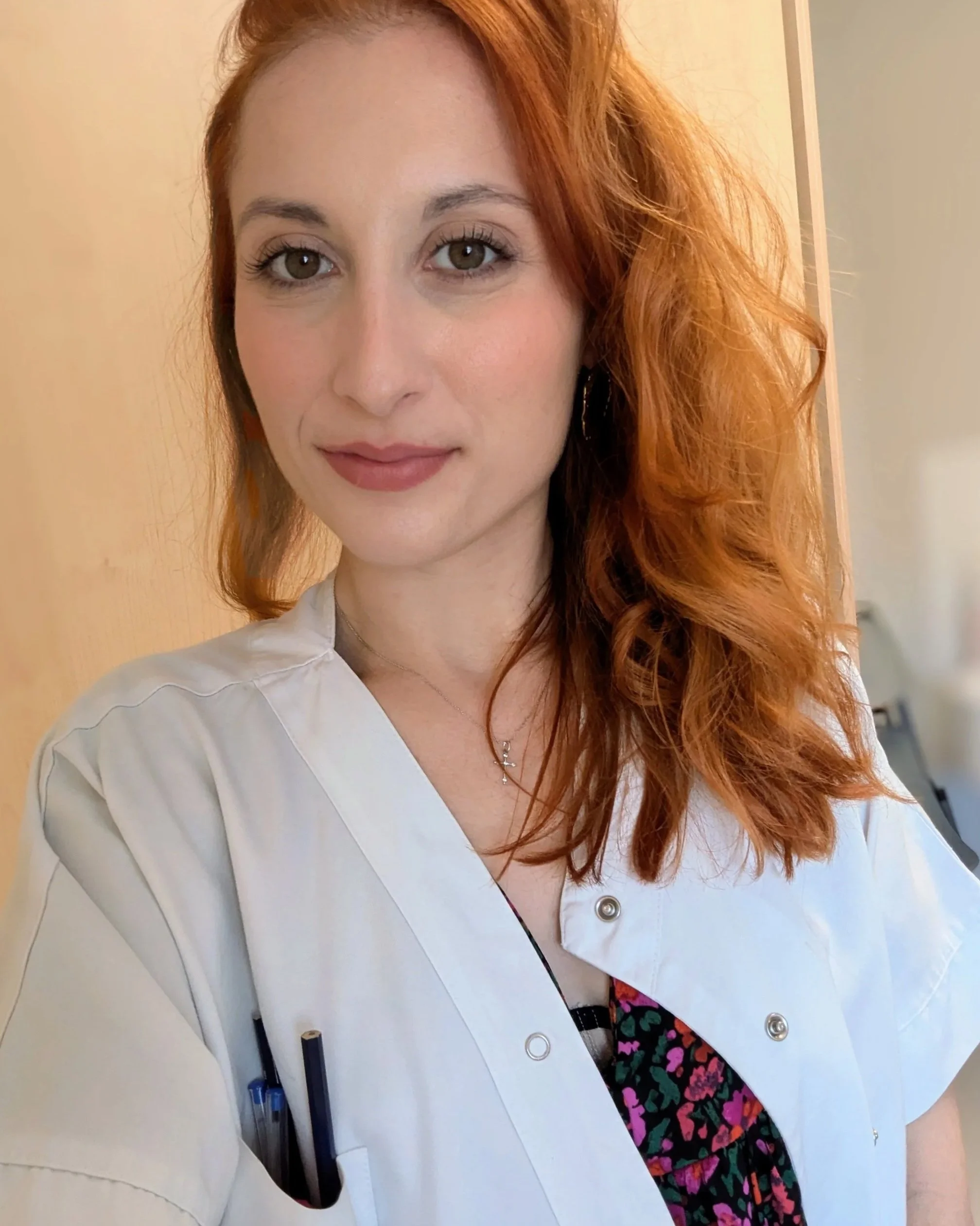 Une femme aux cheveux roux, portant une blouse blanche et un col roulé à motifs colorés, sourit à la caméra dans un intérieur.