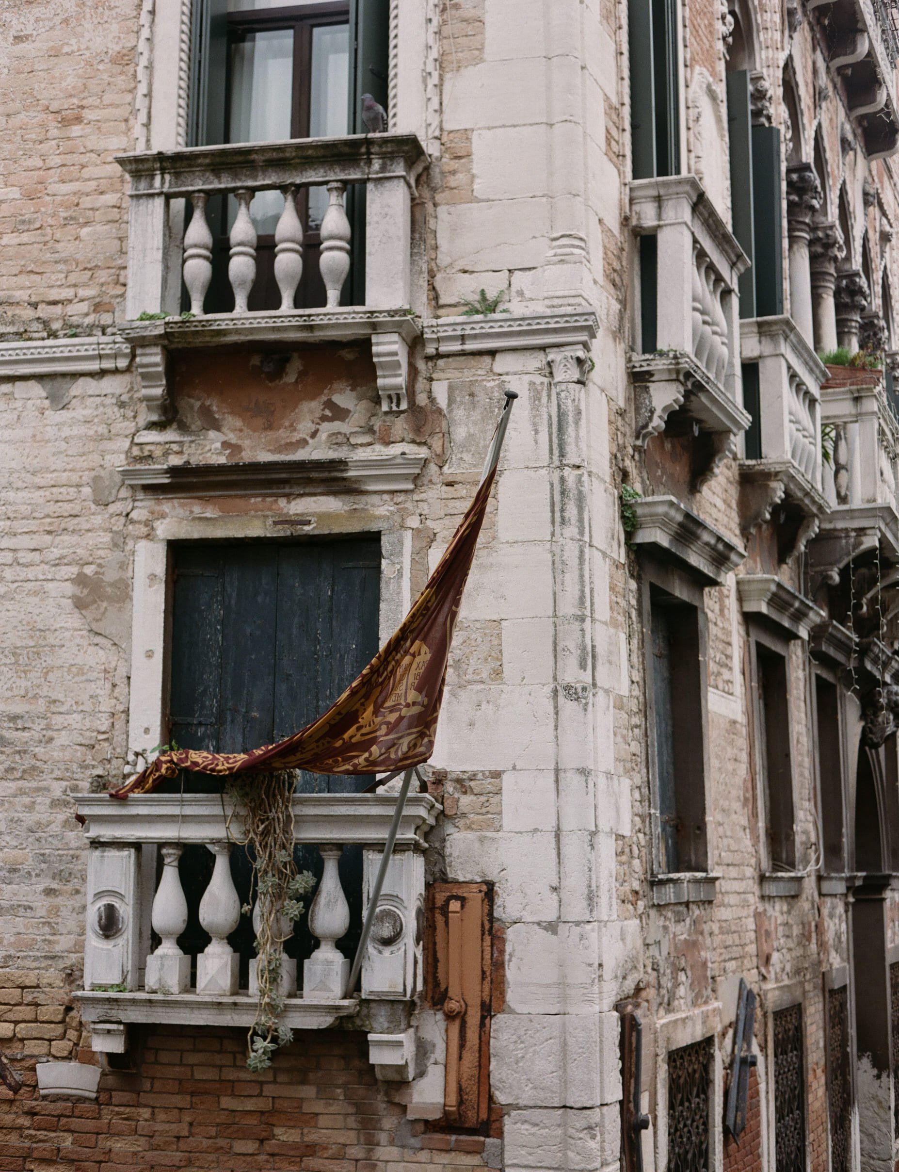 A close-up of an old, weathered building with brick and stone walls, small balconies, and black window shutters. A torn, partially hanging flag is draped over a railing.