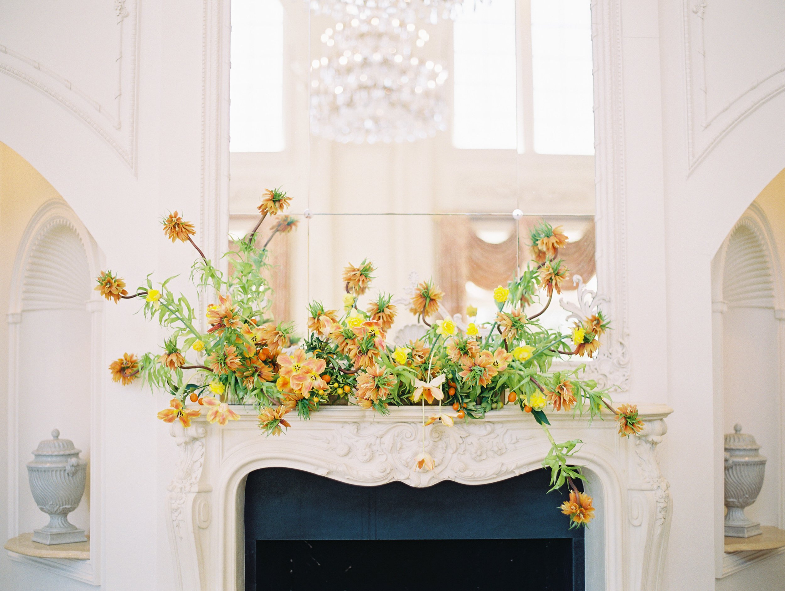 a fireplace mantle with a mirror on top and orange, yellow, and peach flowers across the mantle