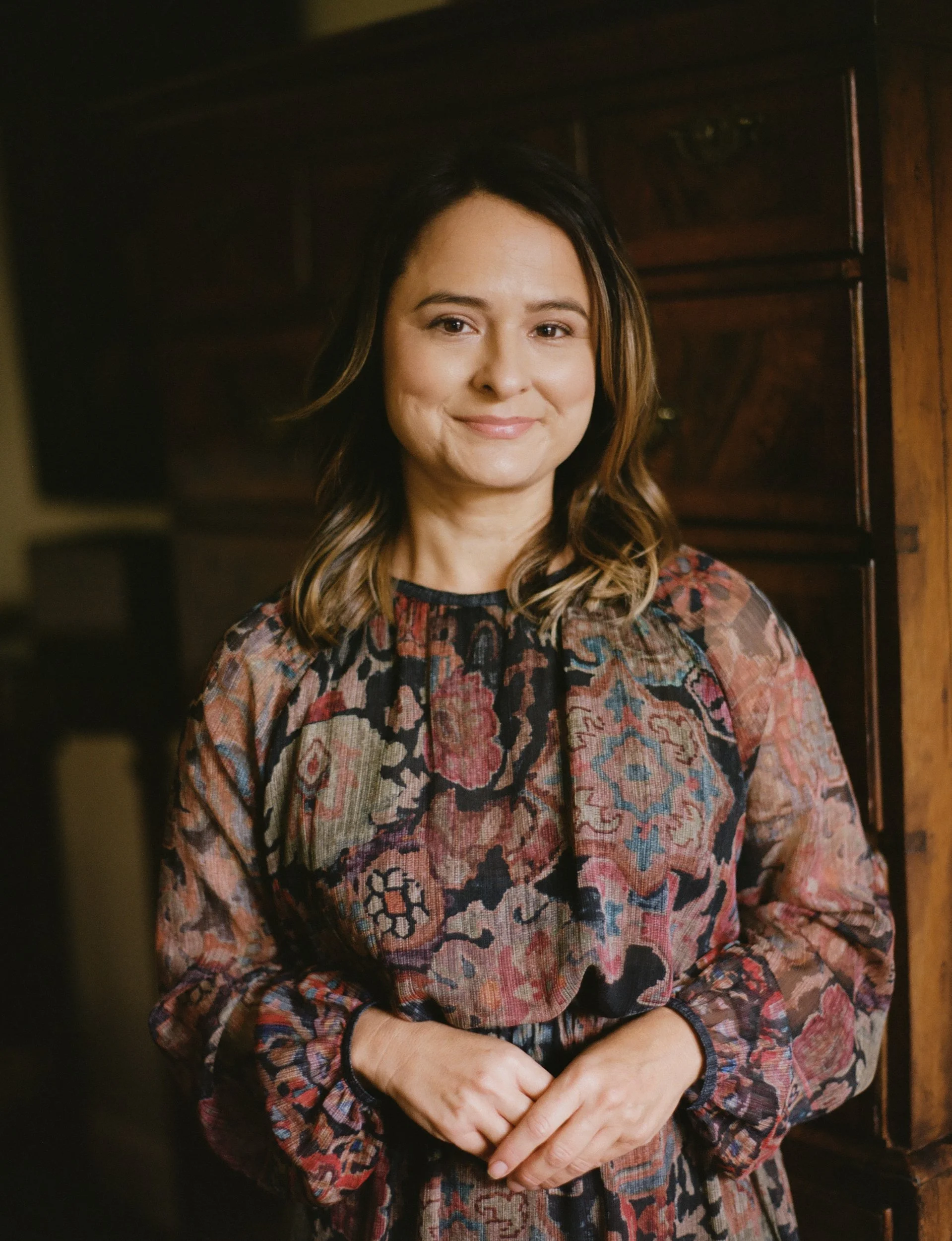 A woman with shoulder-length brown hair wearing a floral patterned dress standing indoors near wooden furniture.