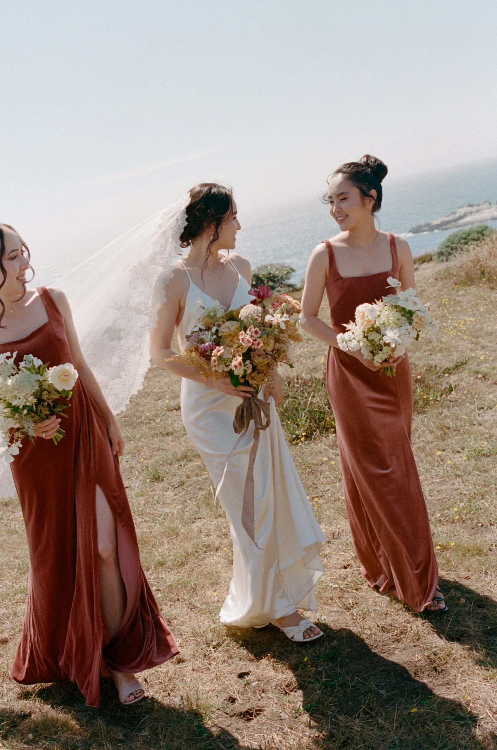 A bride and two bridesmaids walking outdoors on a sunny day near the coast, holding bouquets of flowers, with the sea visible in the background.