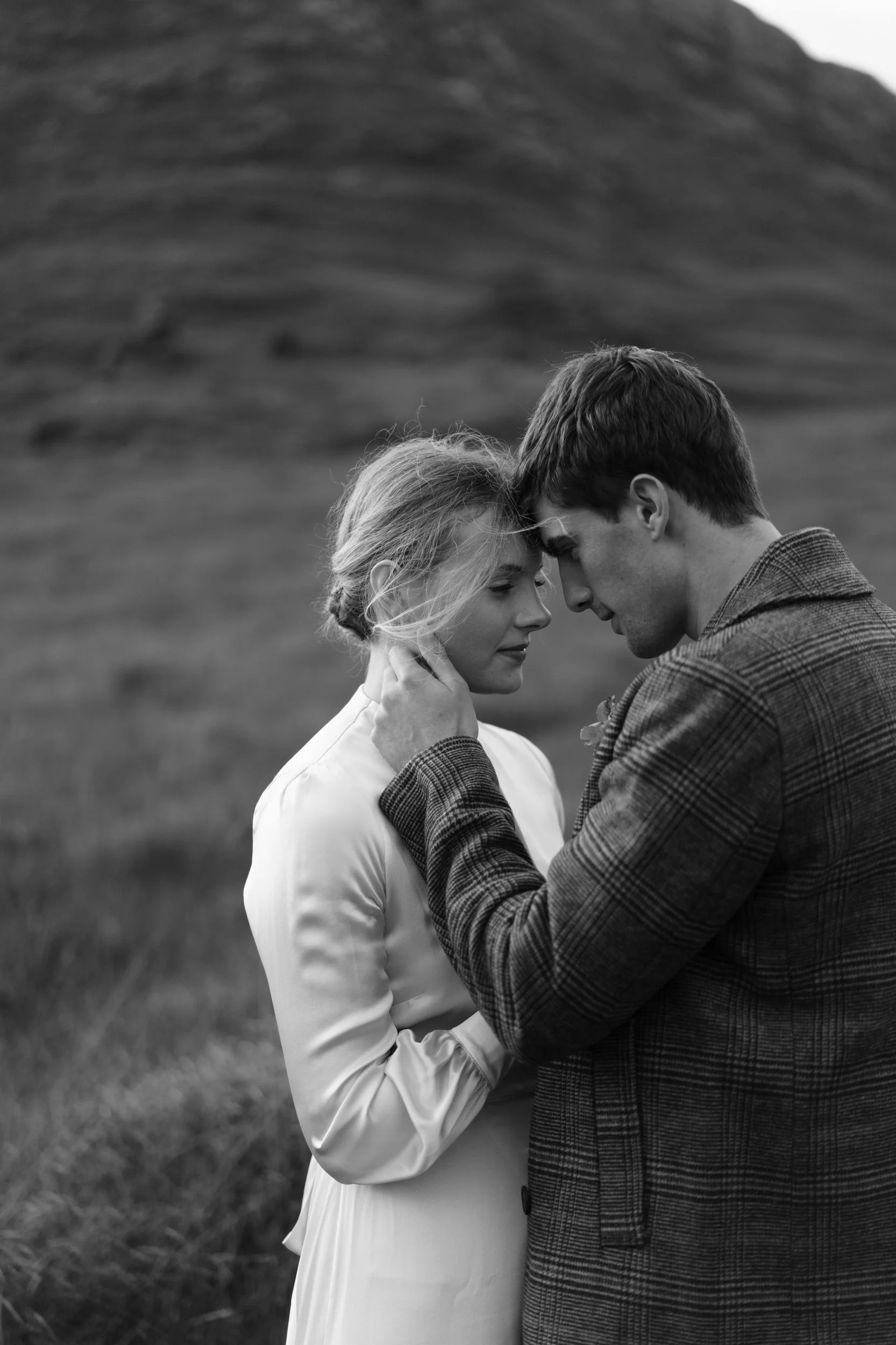 A black and white photo of a man and woman standing close together outdoors, touching foreheads and holding each other's faces in a tender moment, with a mountain or hill in the background.