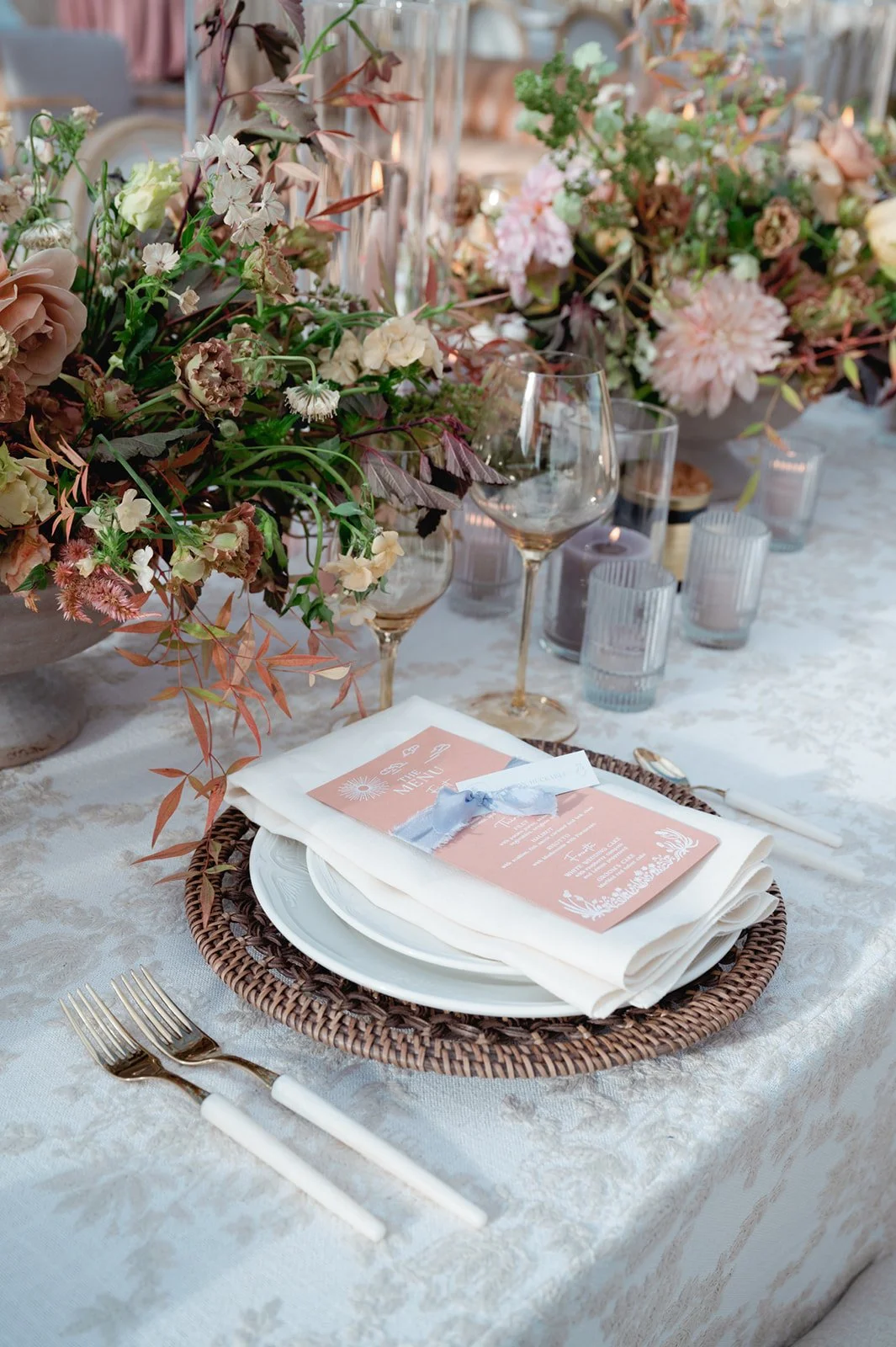 two flower arrangements on a table with a charger and plates and a wine glass and forks in front of it