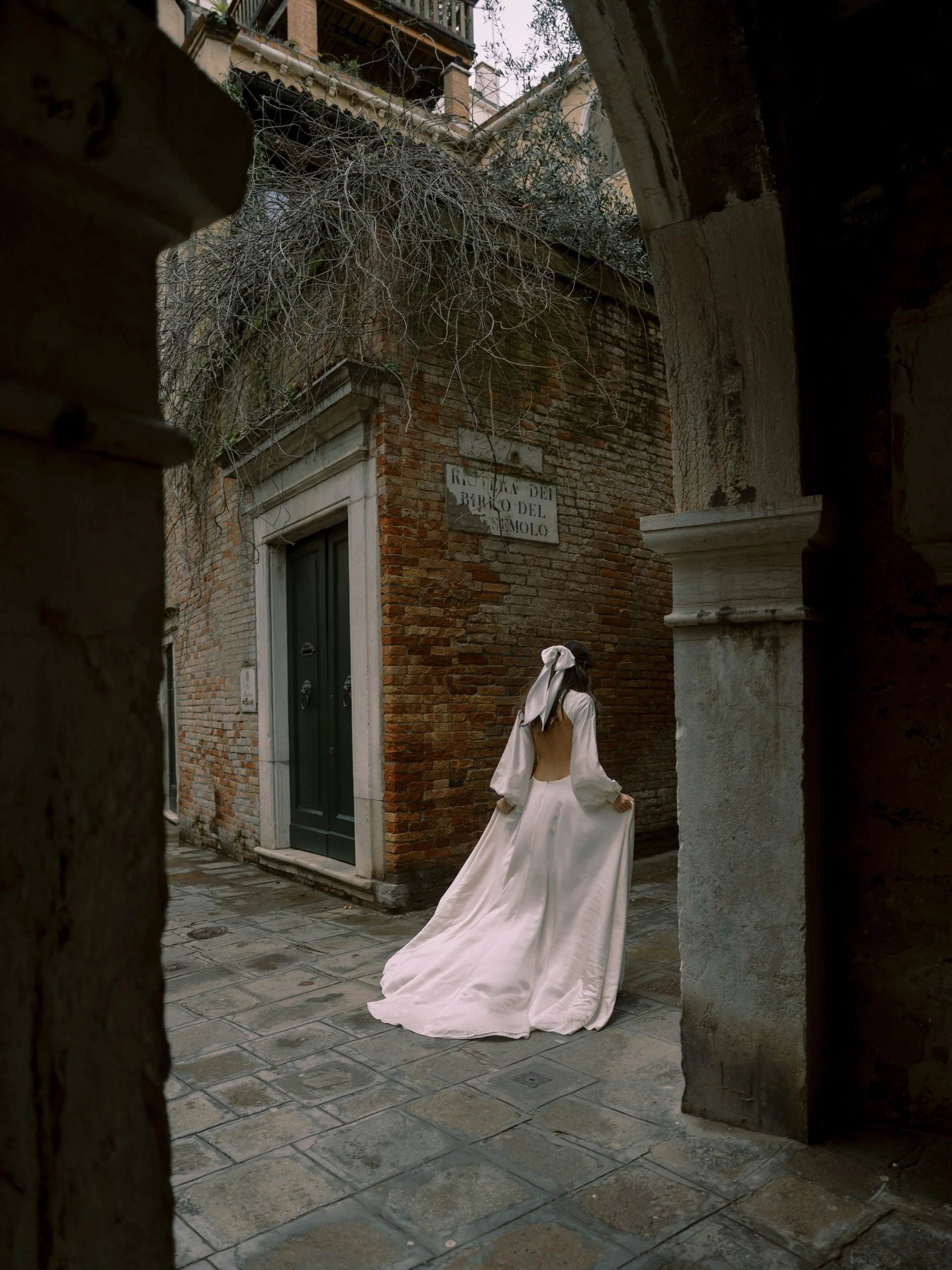 A woman in a long white dress with a bow in her hair standing on a cobblestone street in an alleyway with brick buildings.