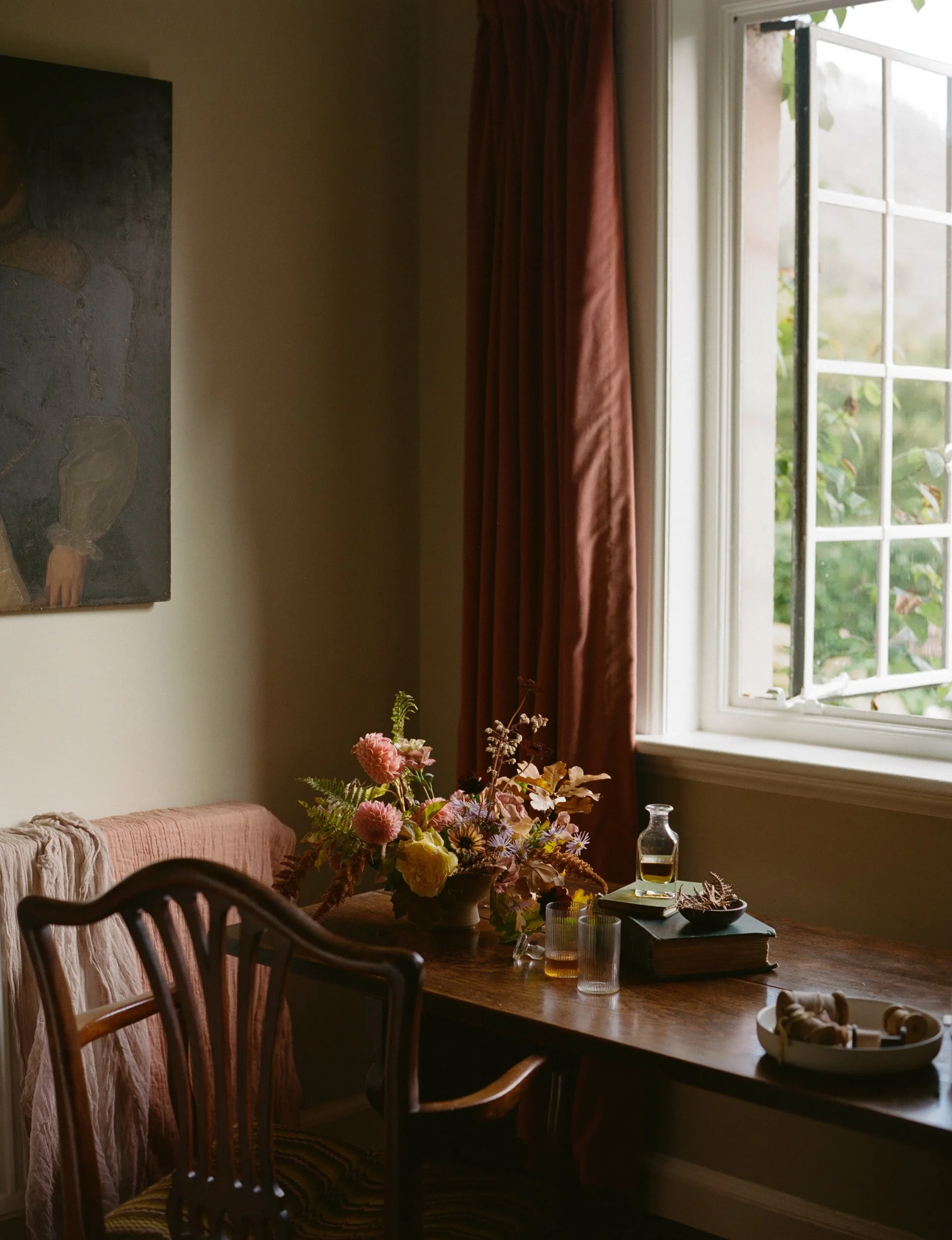 Interior of a room with a wooden table near a window, decorated with a floral arrangement, books, a glass bottle, and small glasses. A pink curtain hangs by the window, and a painting is partially visible on the wall.