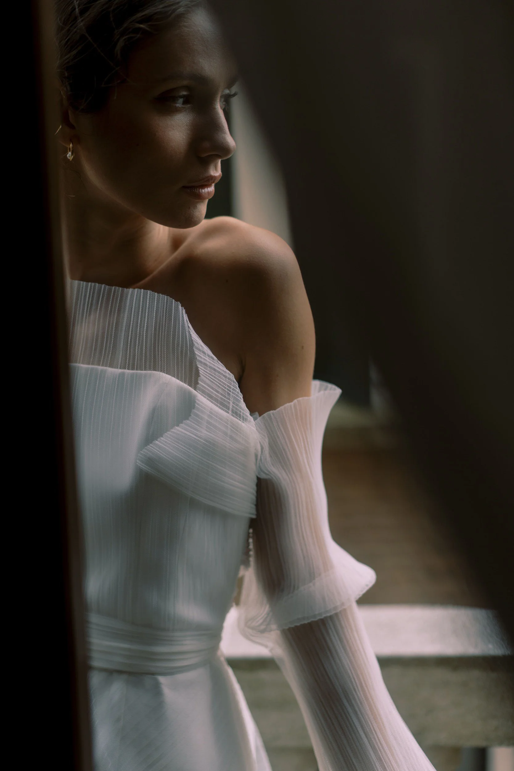 A woman with light skin and curly hair wearing a white off-shoulder dress, gazing out a window with a reflective, thoughtful expression.