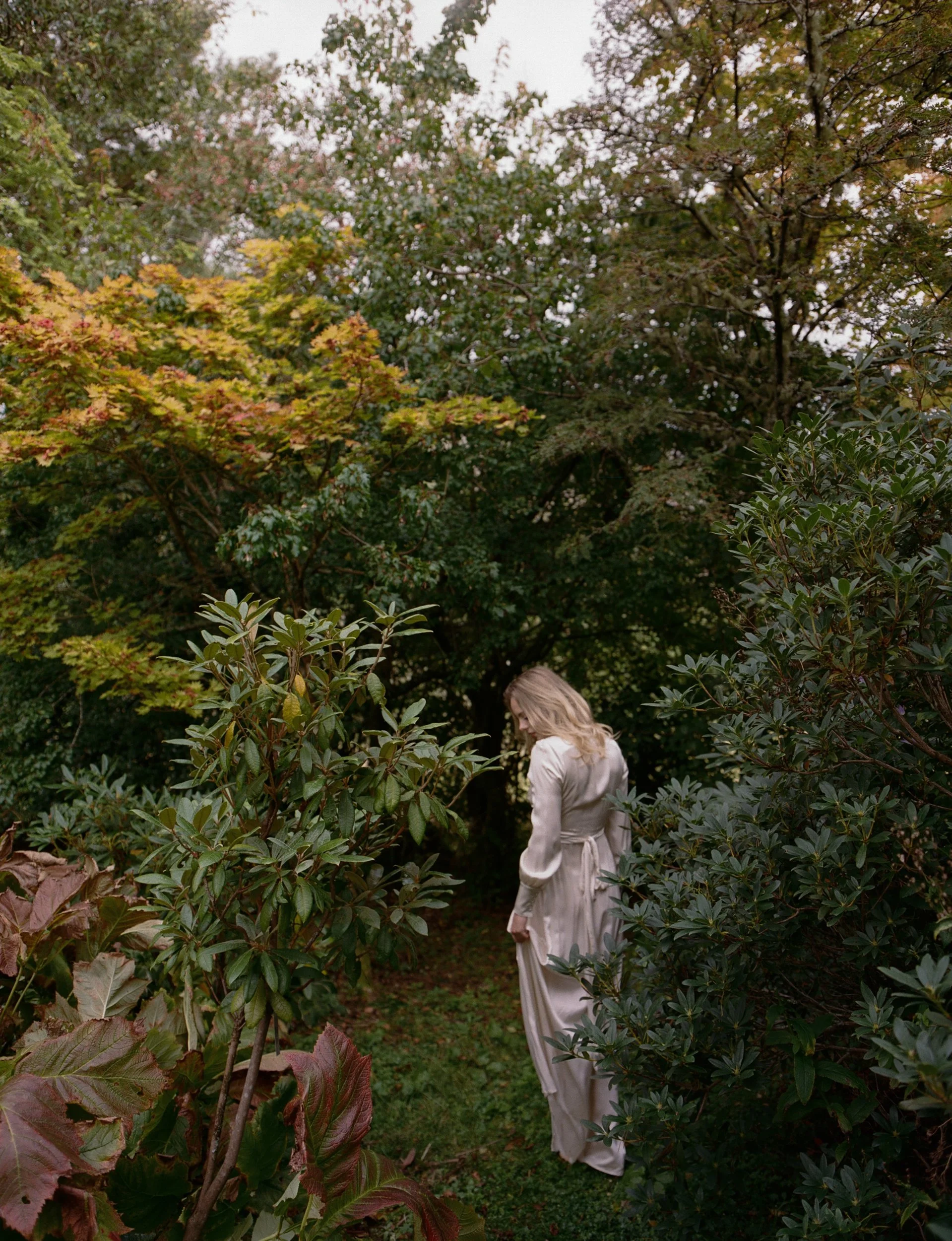 A woman with blonde hair wearing a beige coat and matching dress standing among green and brown trees and bushes in a garden or park.