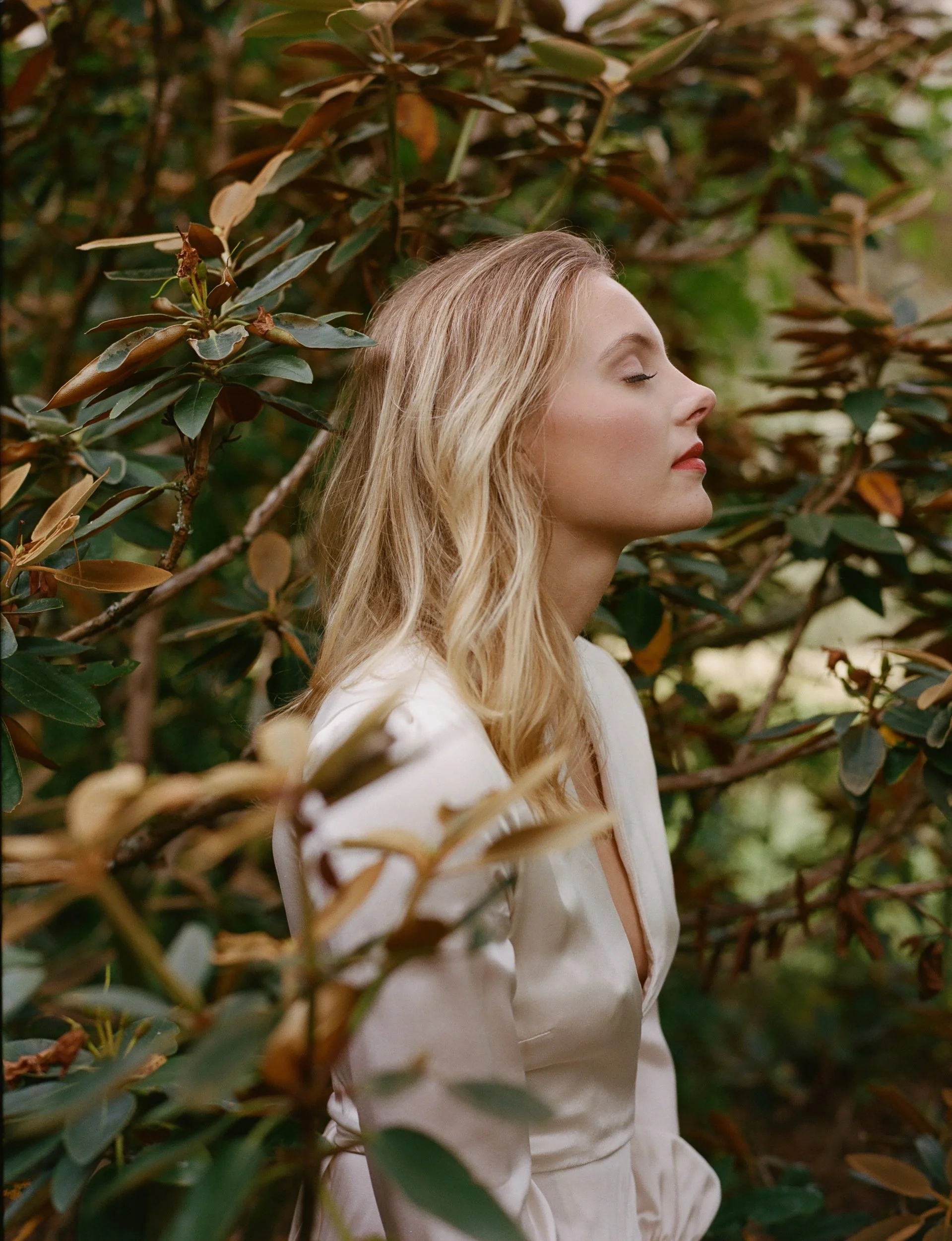 A woman with blonde, wavy hair and fair skin stands with her eyes closed among green and brown leaves, appearing to enjoy the peaceful moment.