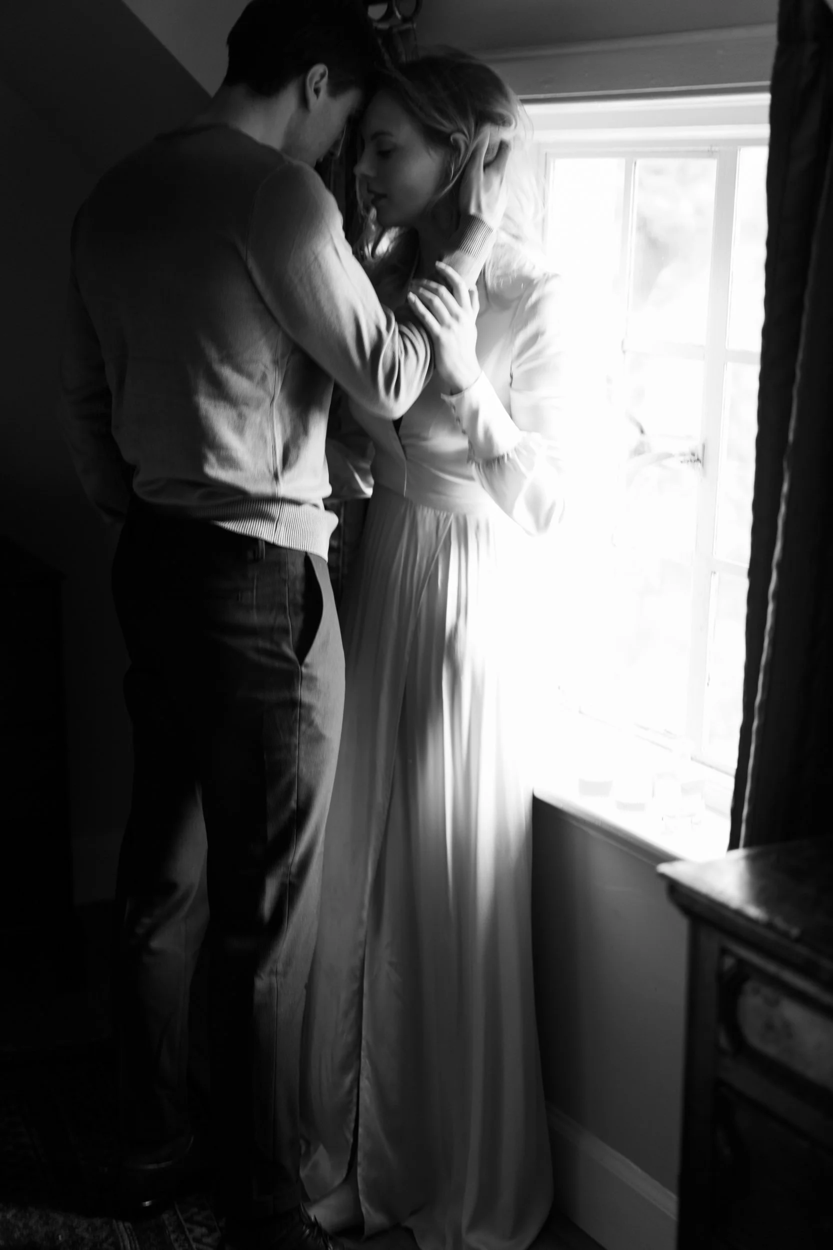 A black and white photo of a couple standing close together near a window, with the sunlight illuminating their faces. The woman is wearing a long dress, and the man is in casual clothes. They are touching foreheads and engaging in an intimate moment.