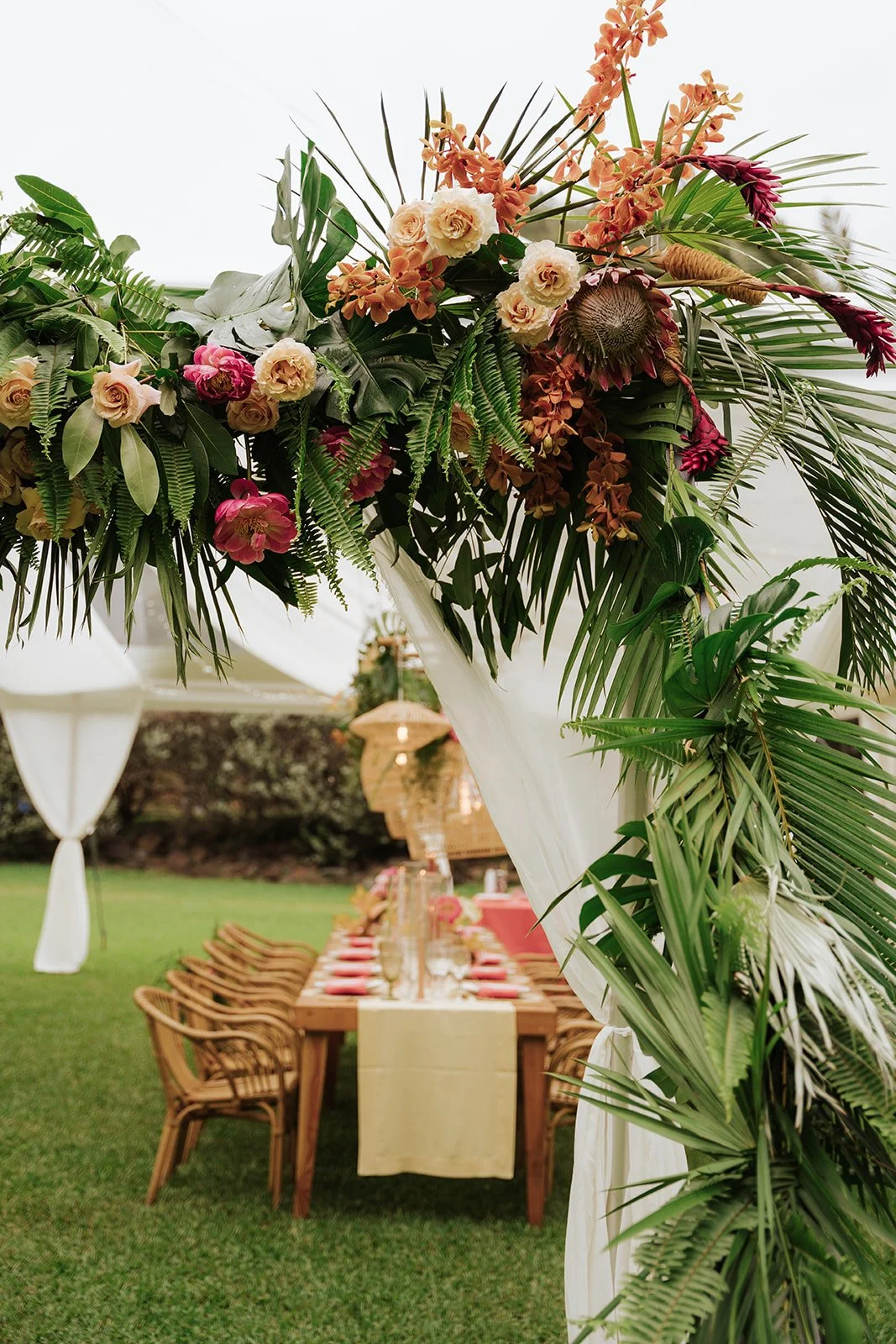 tropical greens and pink flowers lining white drapery with a dinner table behind